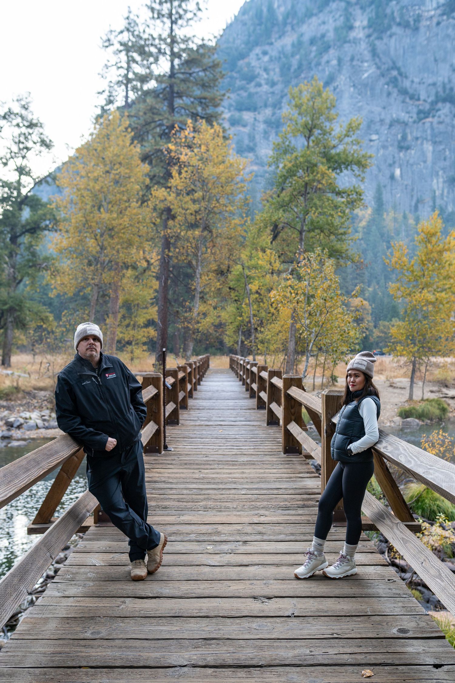 Two people pose casually on a wooden bridge surrounded by autumn trees and mountain scenery.