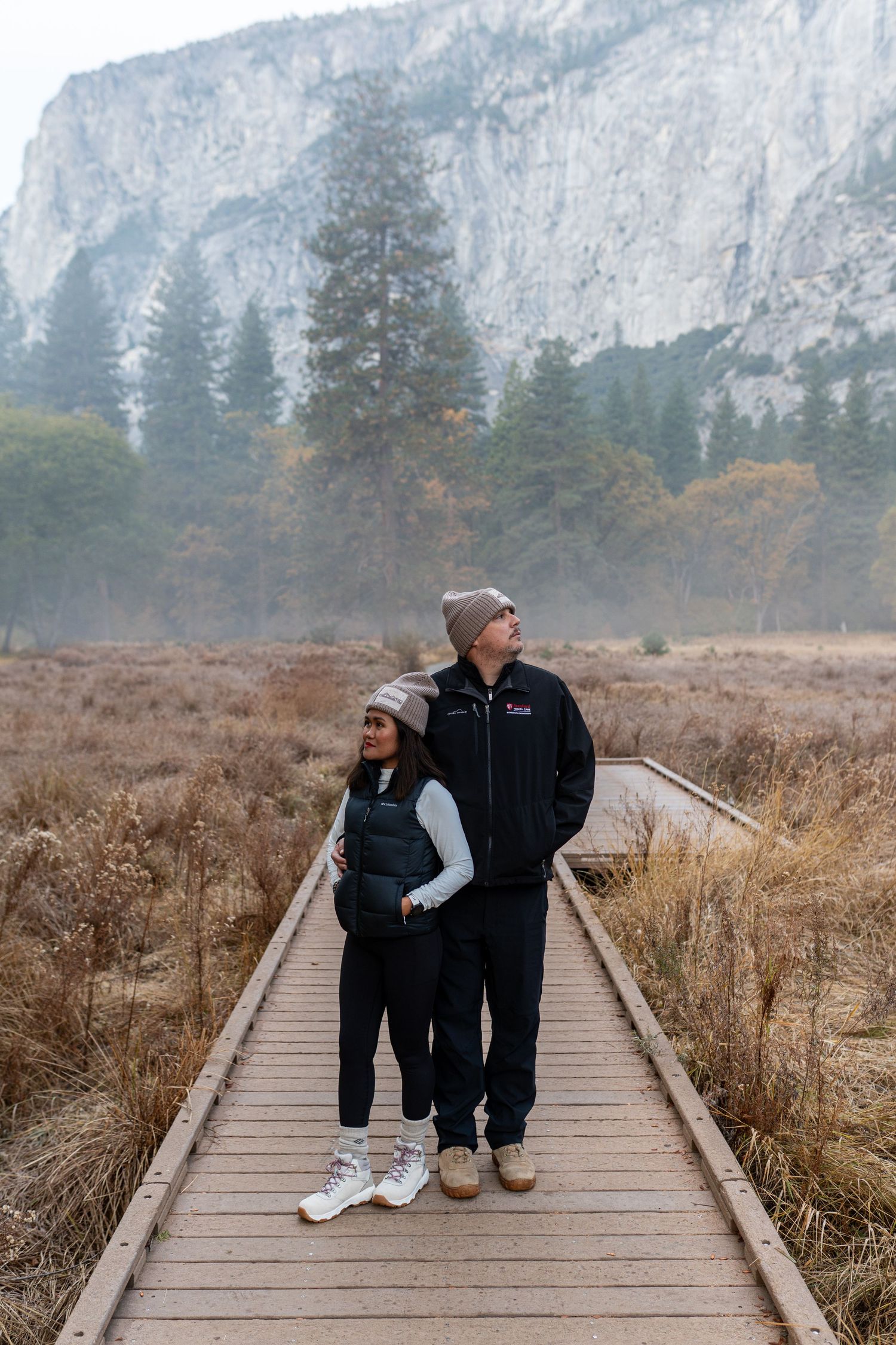 Two people share a moment on a wooden walkway surrounded by tall grass and mountain scenery.