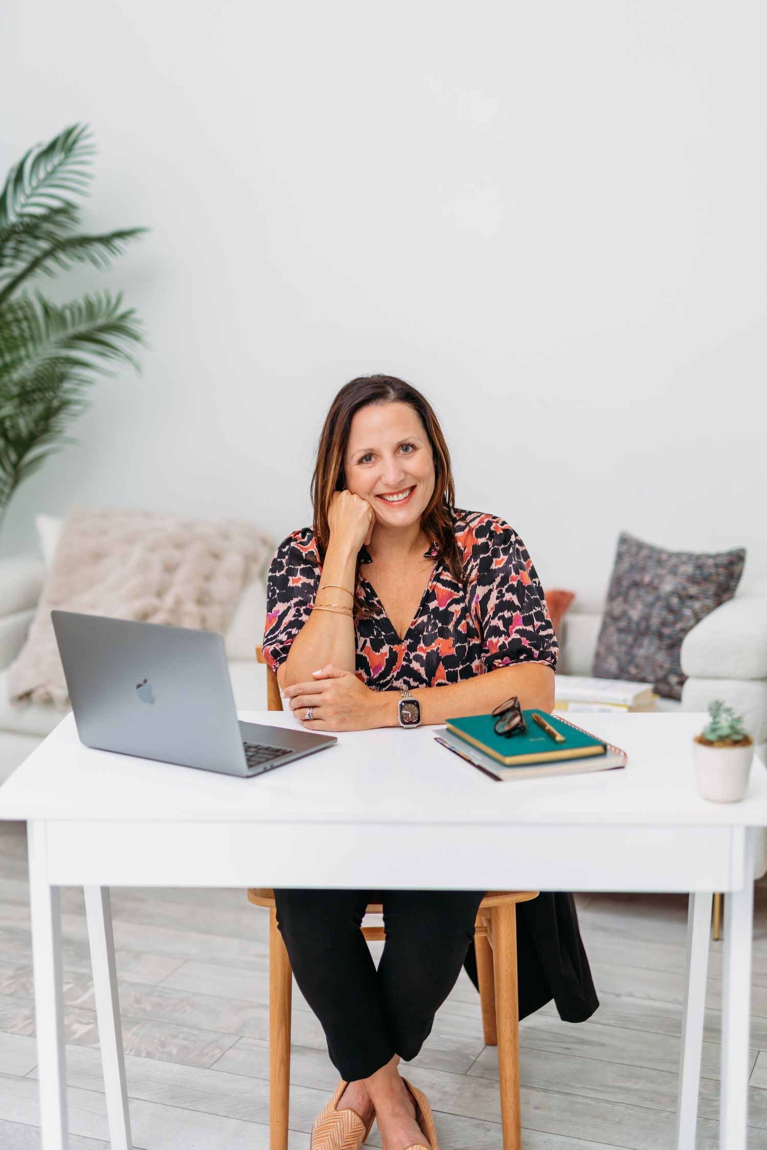 Professional working at white desk with laptop, books and plants in bright modern home office setting.
