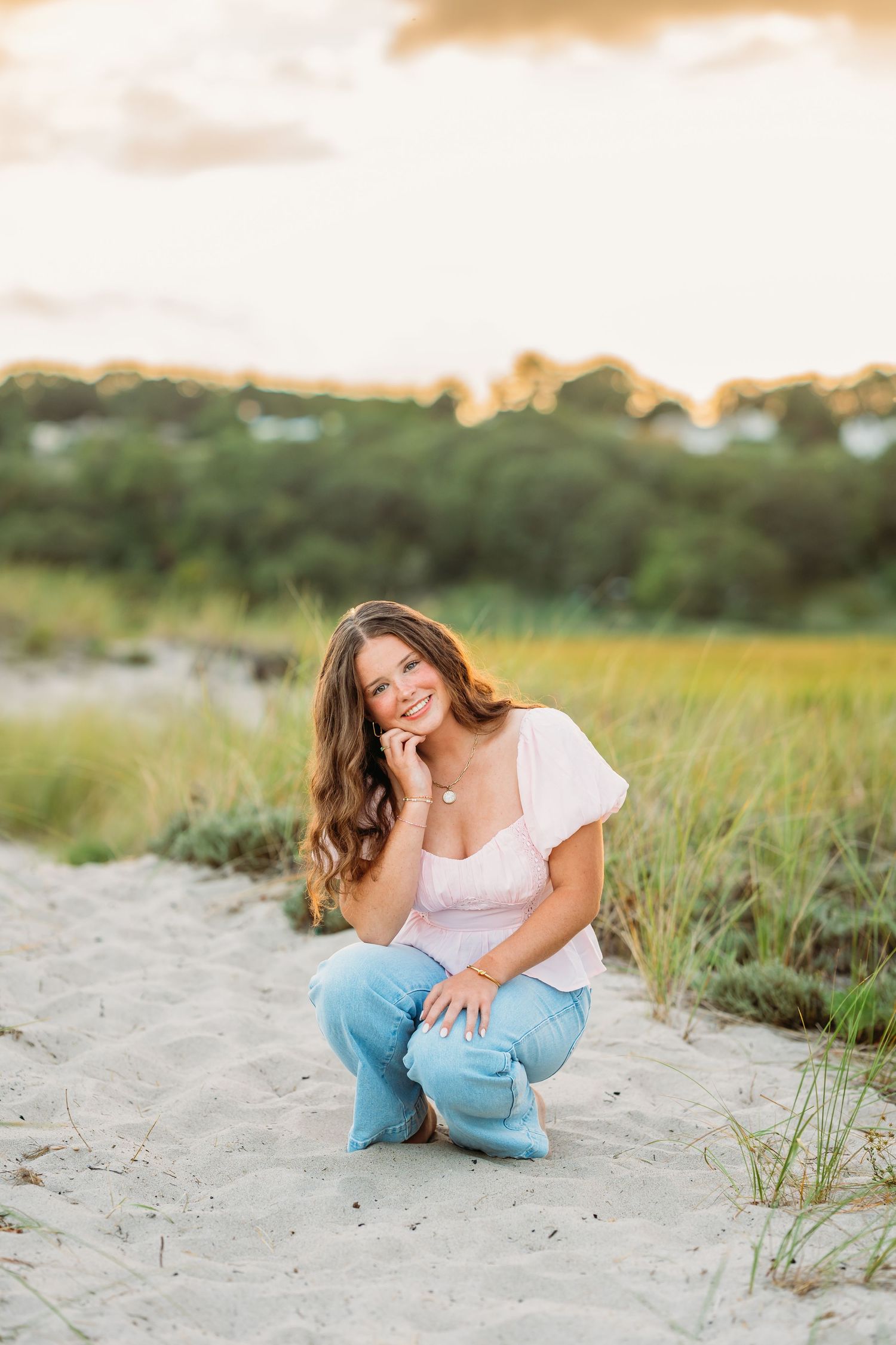 A person in a white top and blue jeans crouches on a sandy beach path at sunset with coastal grass in the background.