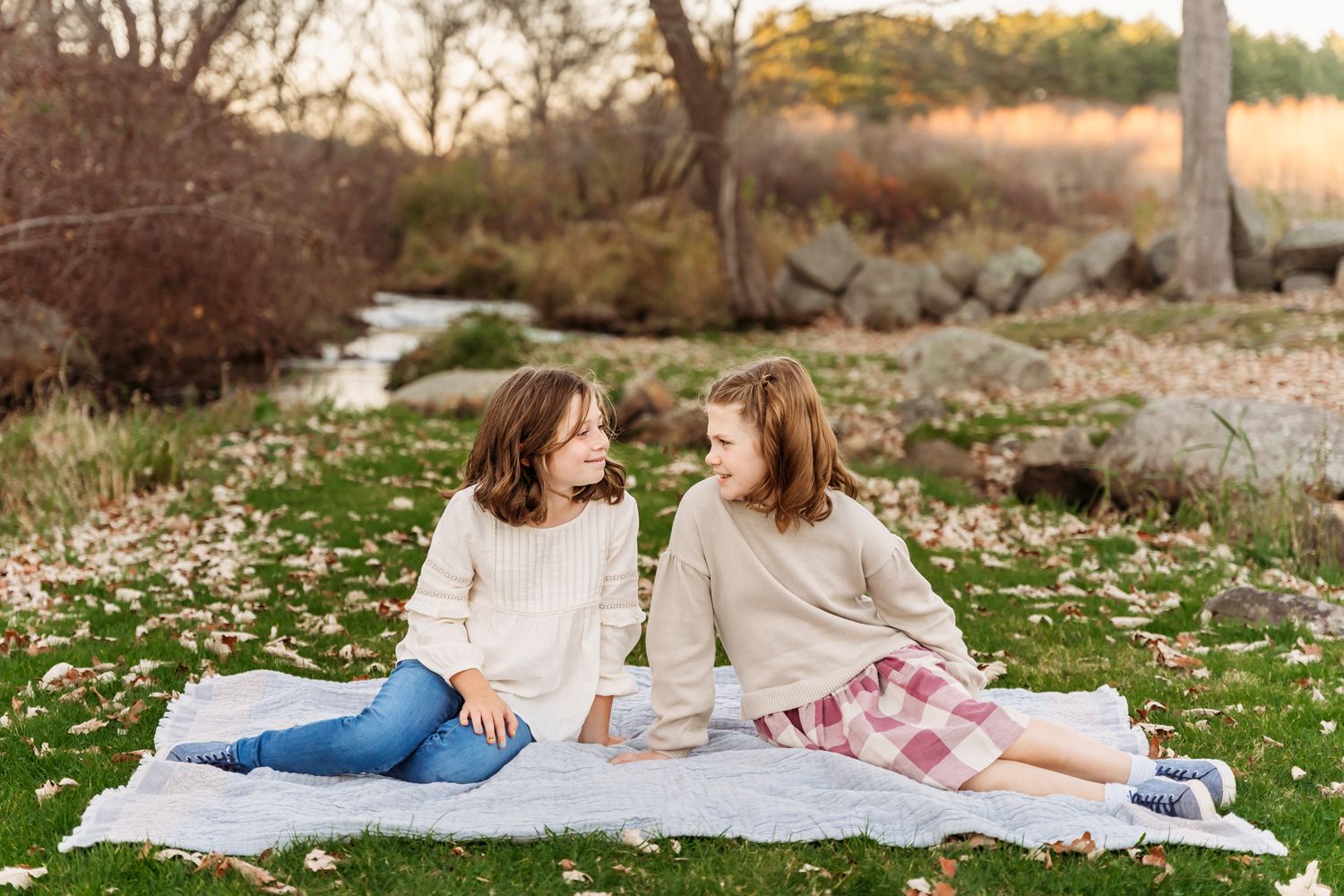 Two people sit on a picnic blanket in autumn with fallen leaves and a stream in the background.