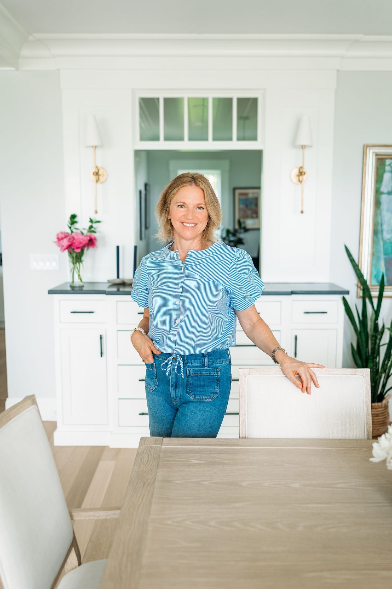A person in denim and blue top sits casually at a modern white dining table in a bright, airy home interior.