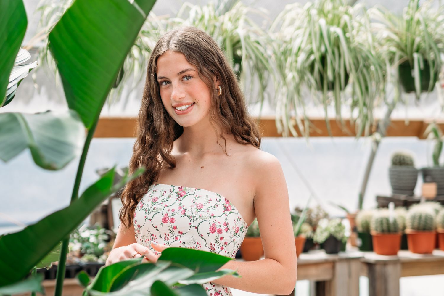 Young woman in floral strapless top smiling among tropical plants and cacti in a greenhouse.
