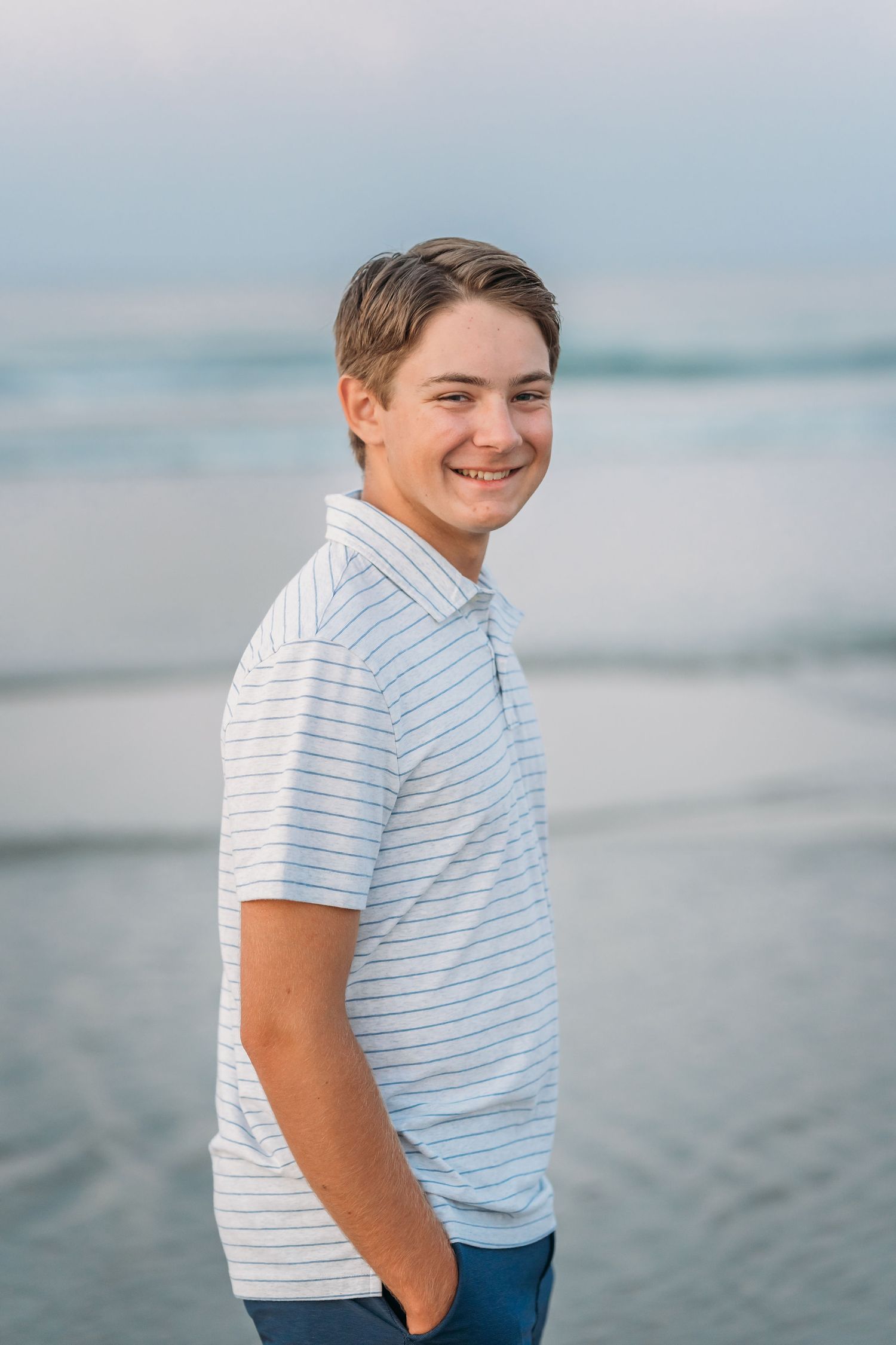 Smiling teenage boy in striped polo shirt standing on beach with ocean waves in background.