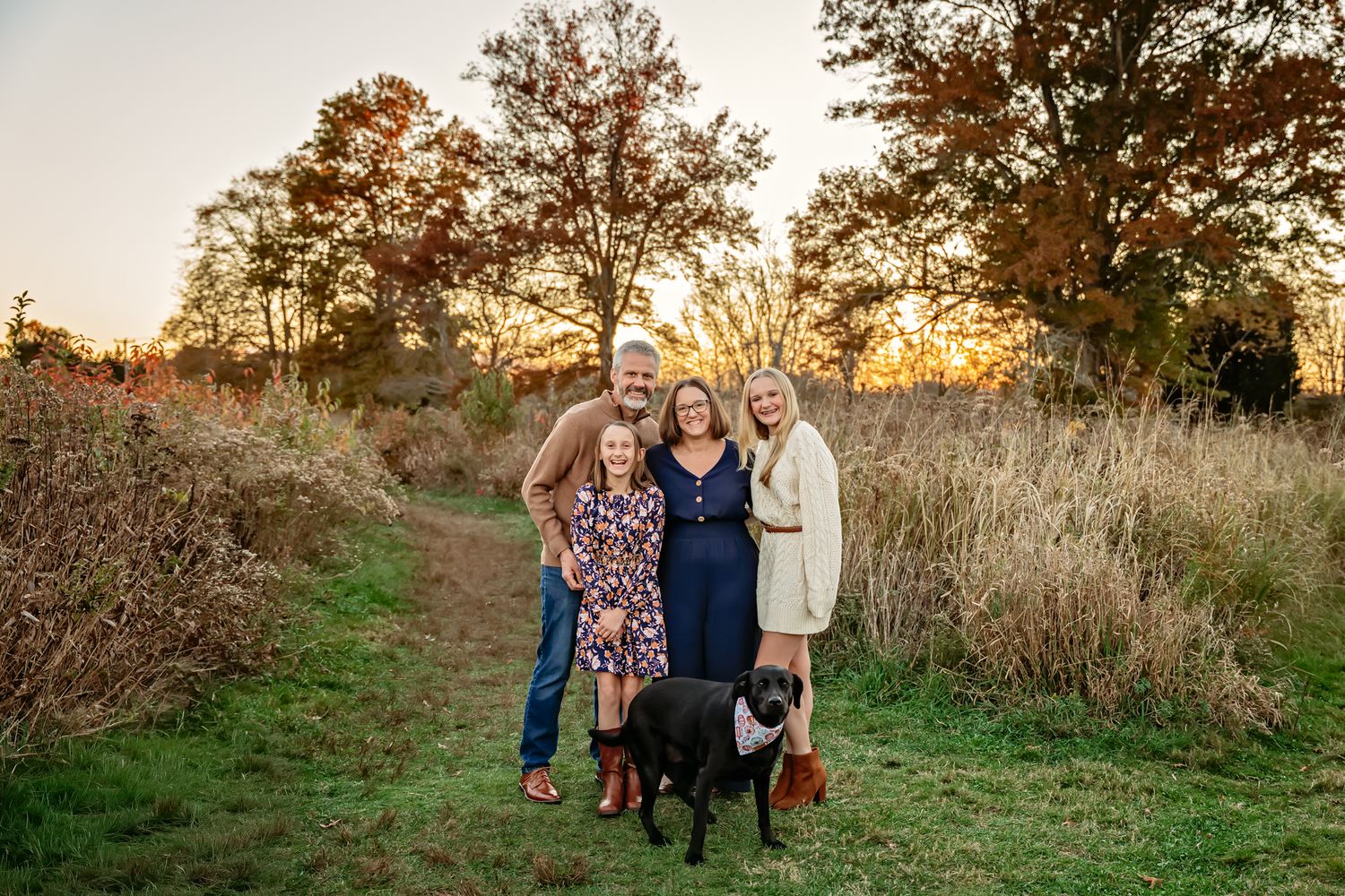 Family of four poses with black dog at golden hour in autumn field with trees and warm sunset light.