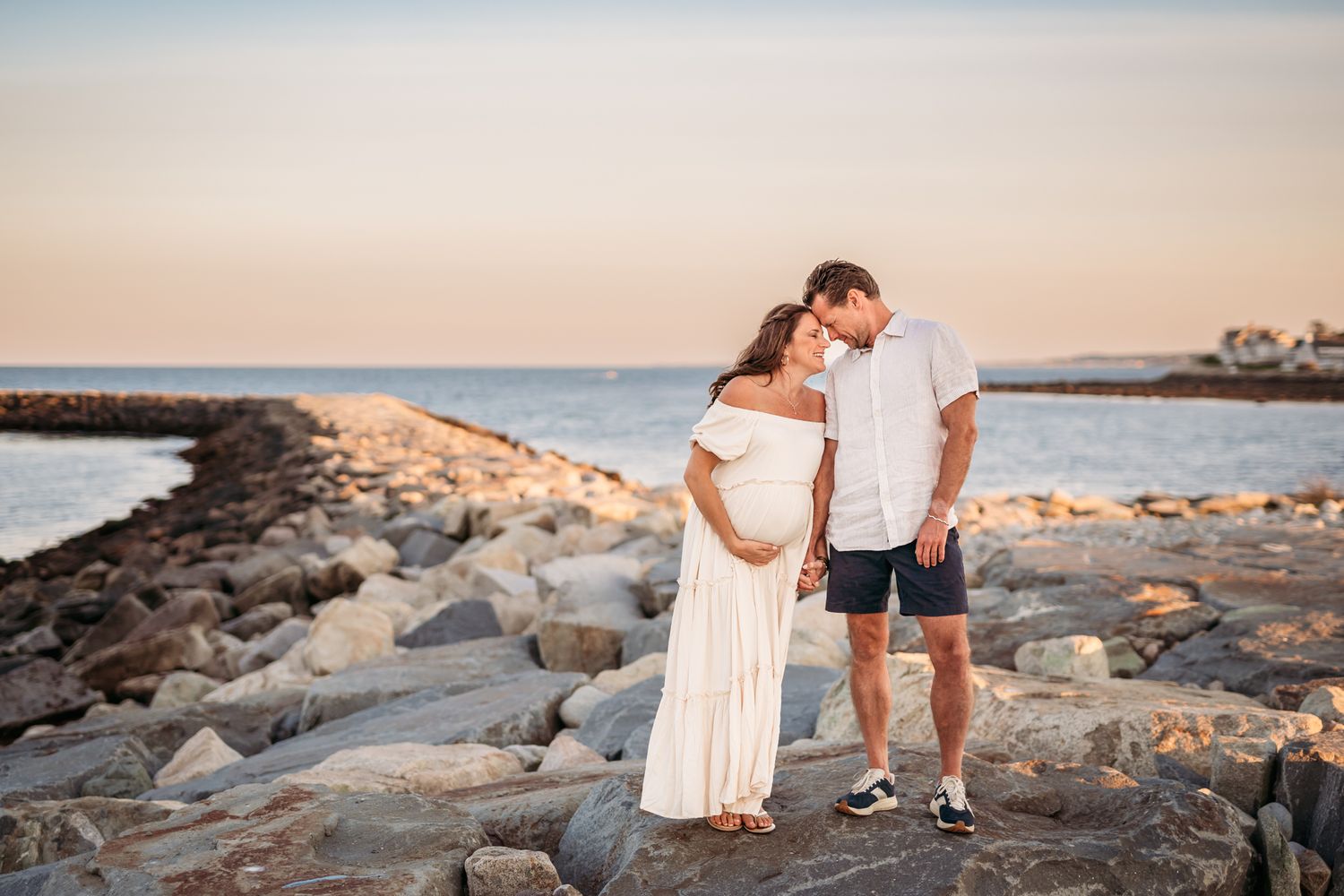 Couple stands together on rocky coastal outcrop during golden hour.