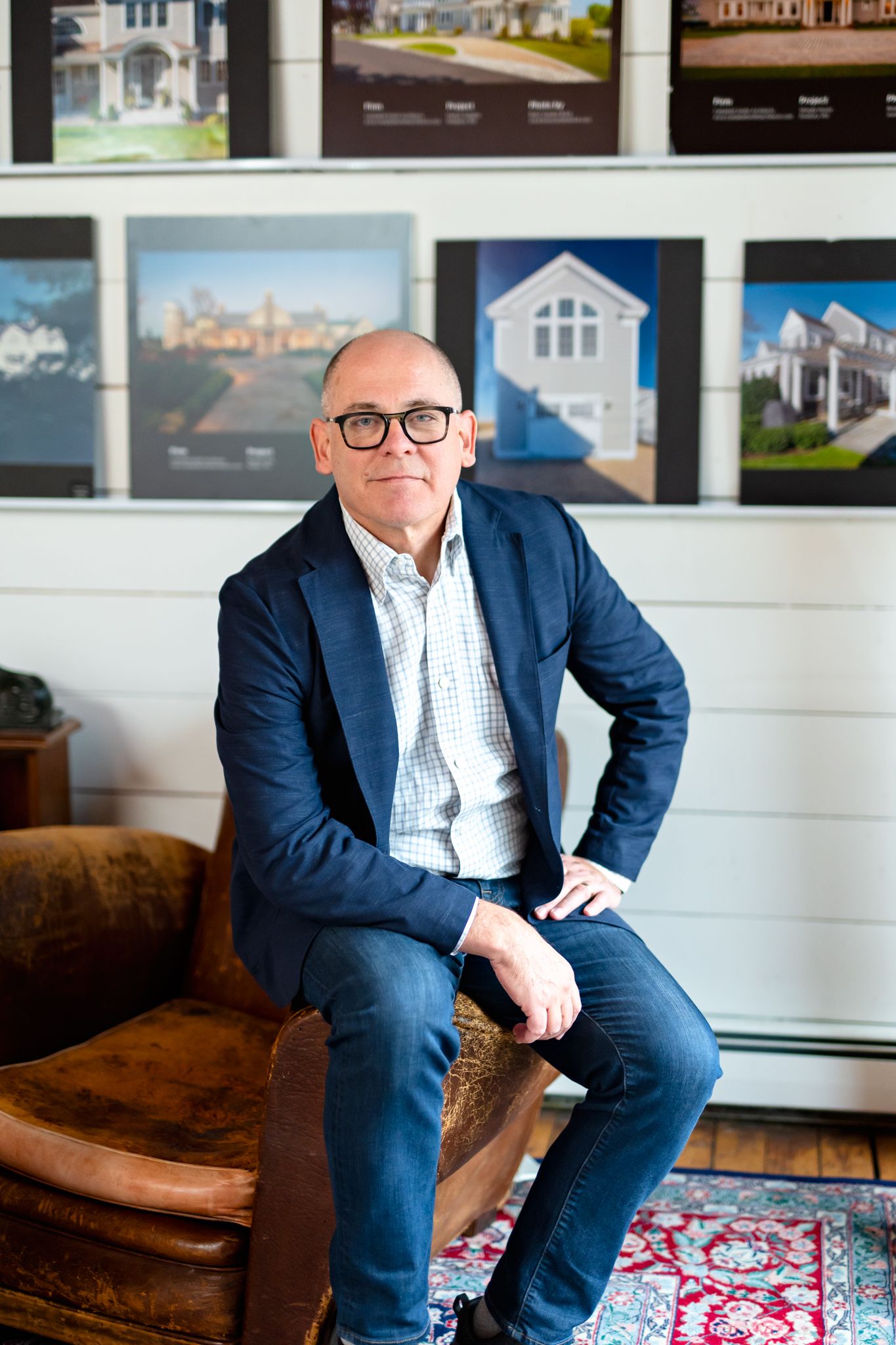 A bald man in glasses and navy blazer sits in a leather chair before a wall of architectural photos.