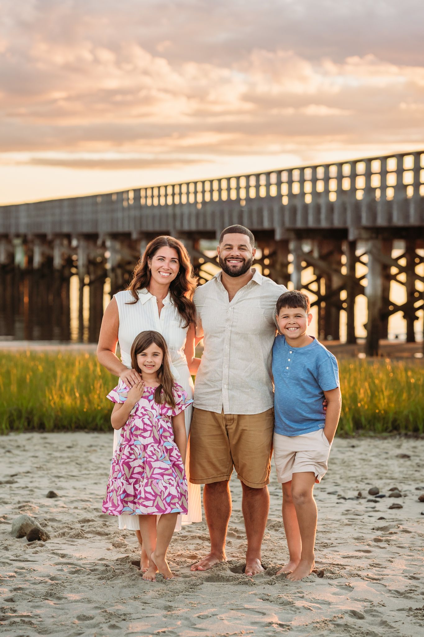 Family of four smiling on sandy beach at sunset with wooden pier in background.