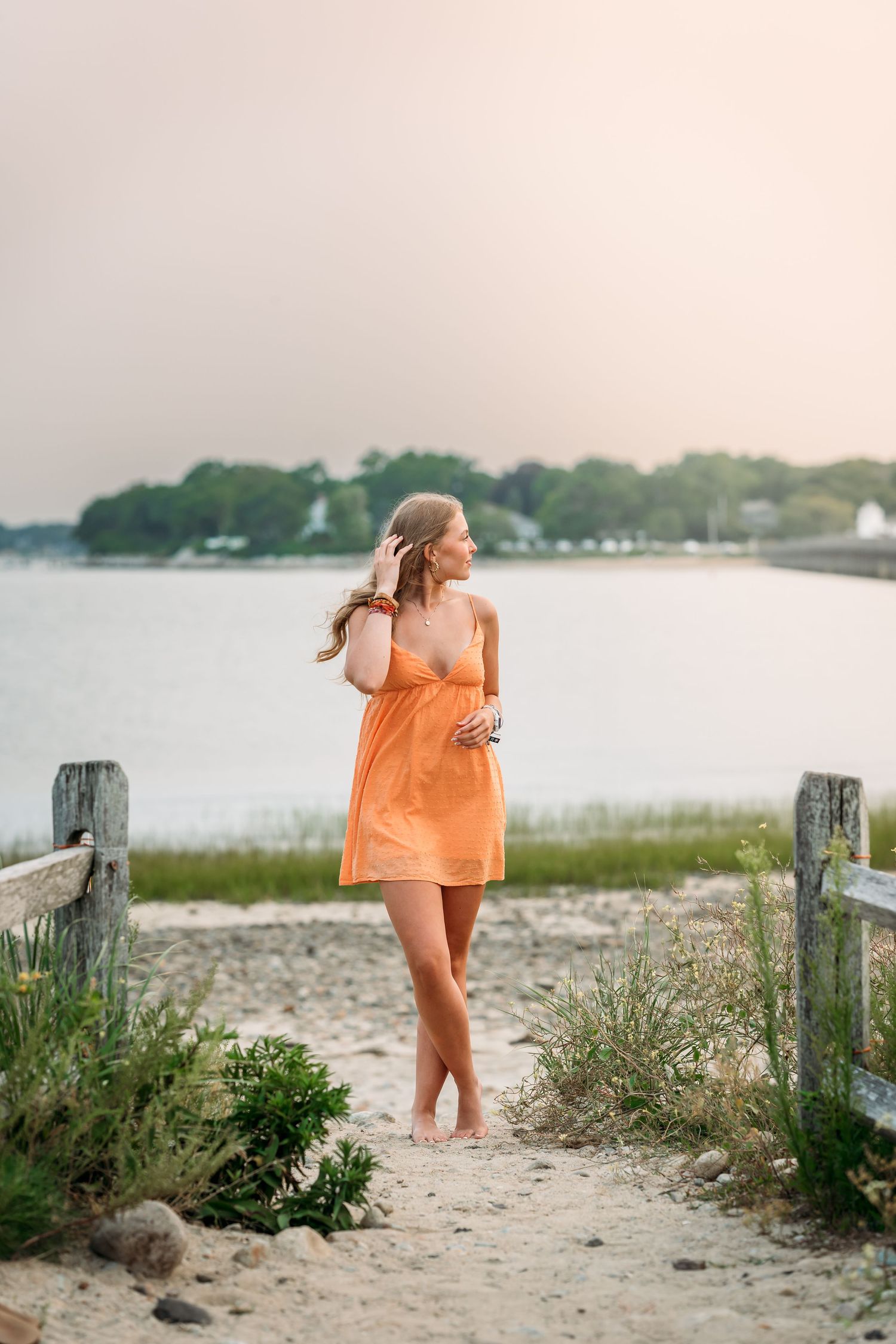 A barefoot woman in an orange sundress stands on a sandy beach path at sunset, with calm water and trees in the background.