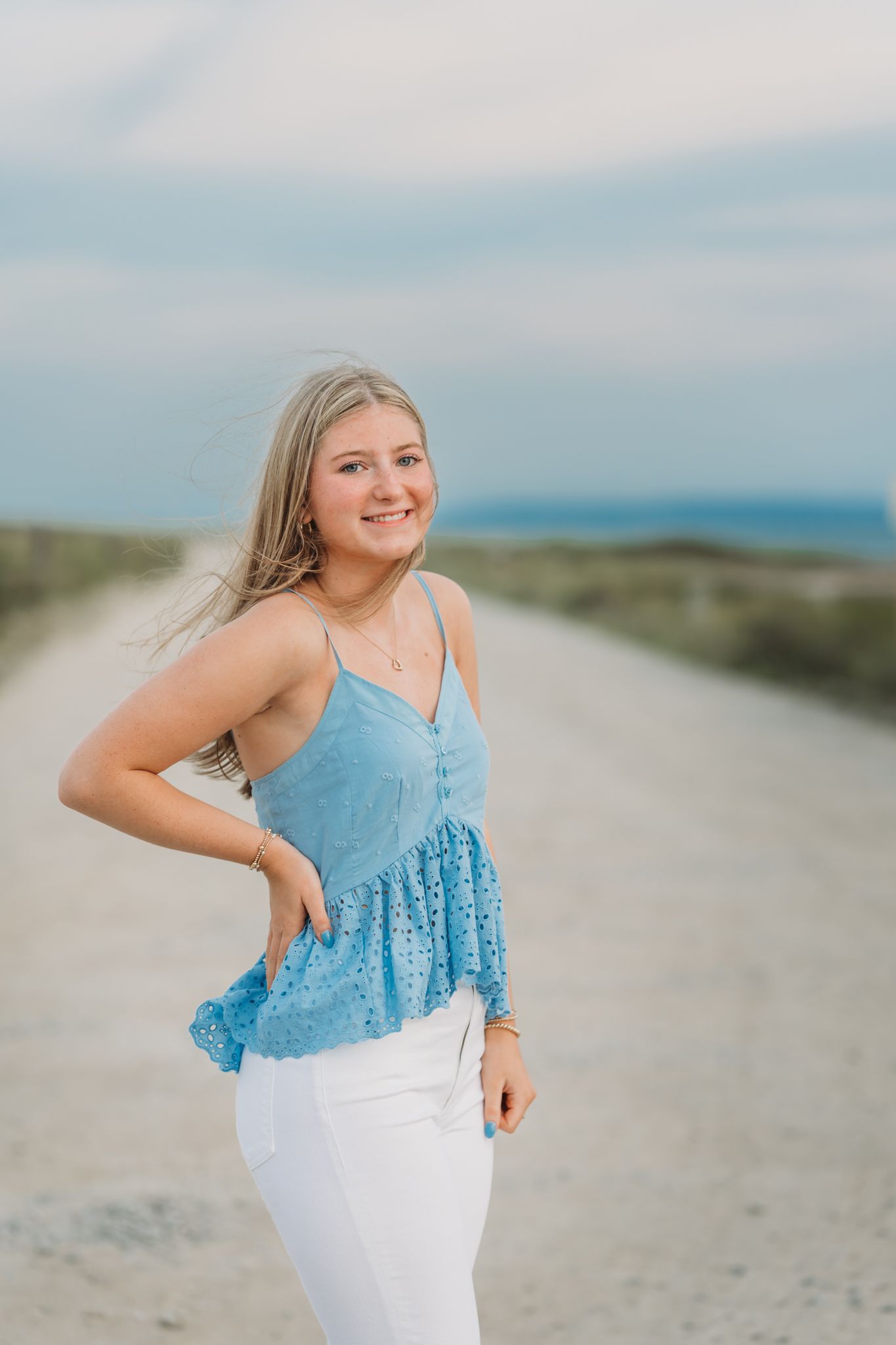 Young blonde woman in blue eyelet top and white jeans smiling on a sandy coastal path.
