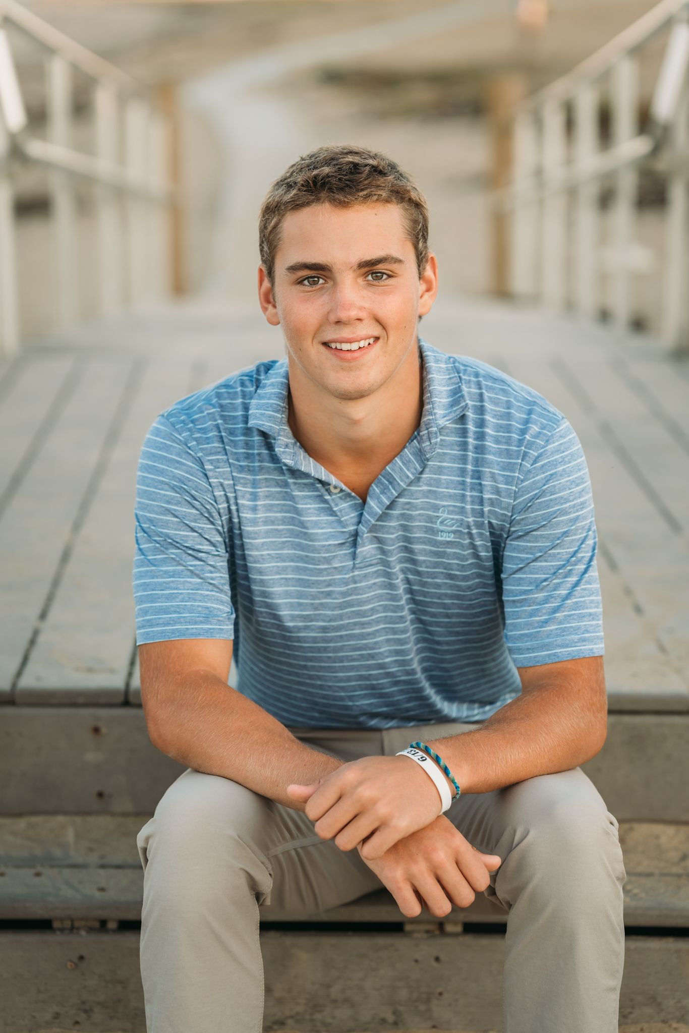 Young man in blue striped polo sits on wooden pier steps, smiling confidently in a senior portrait photo.