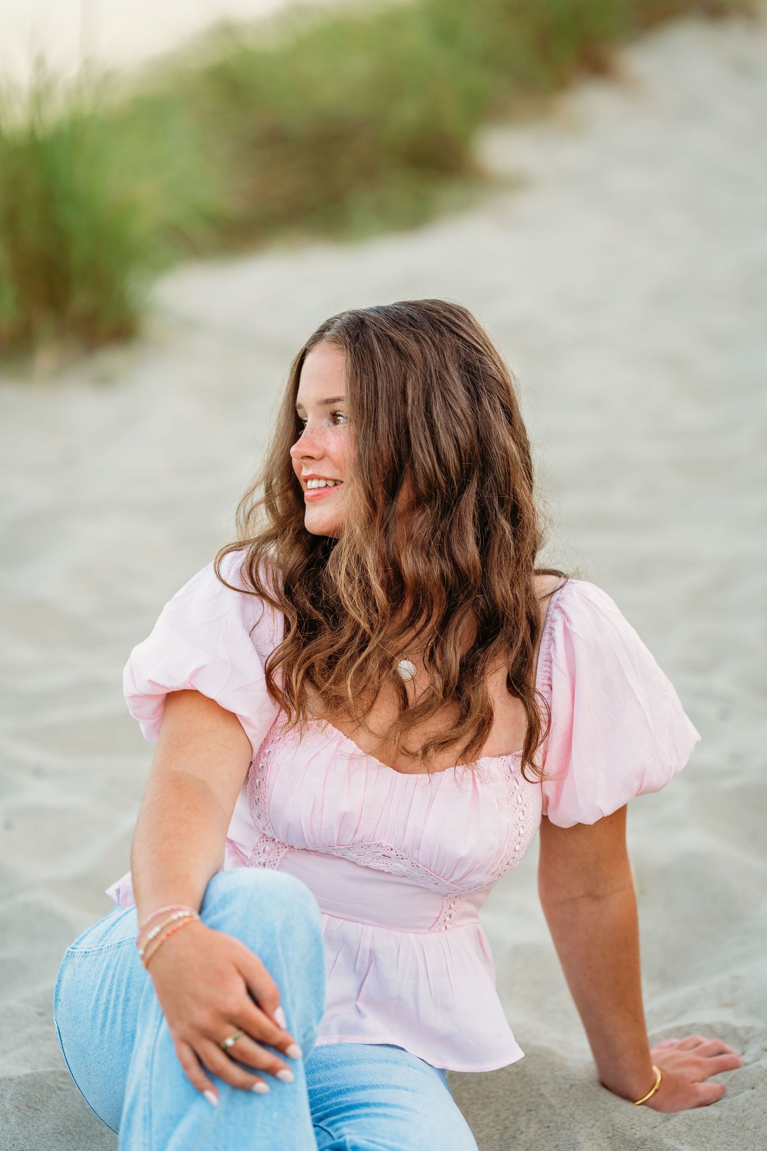 A person in a pink top and blue pants sits casually on a sandy beach with natural greenery in the background.