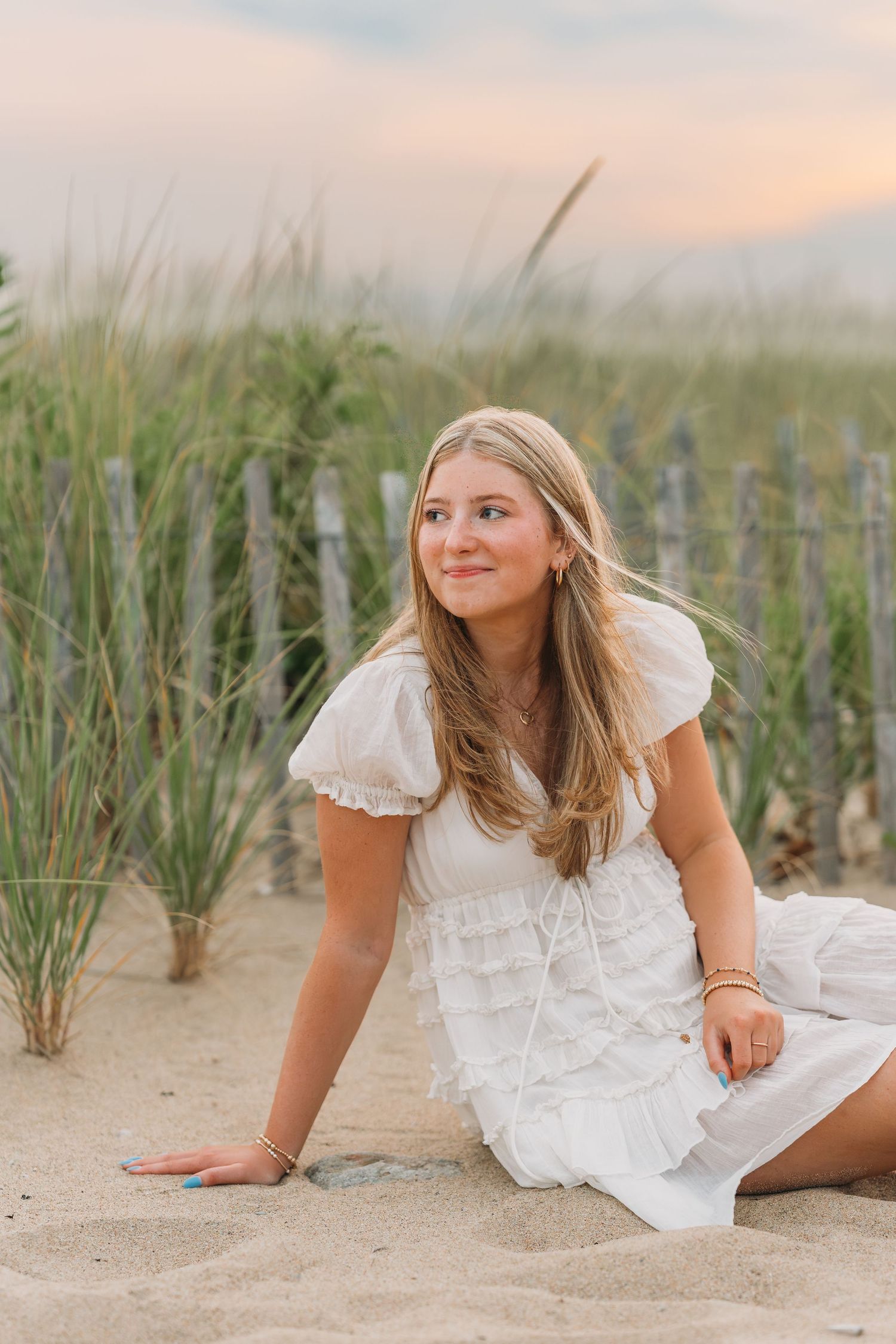 Young woman in white dress sitting on beach sand near dunes at sunset.