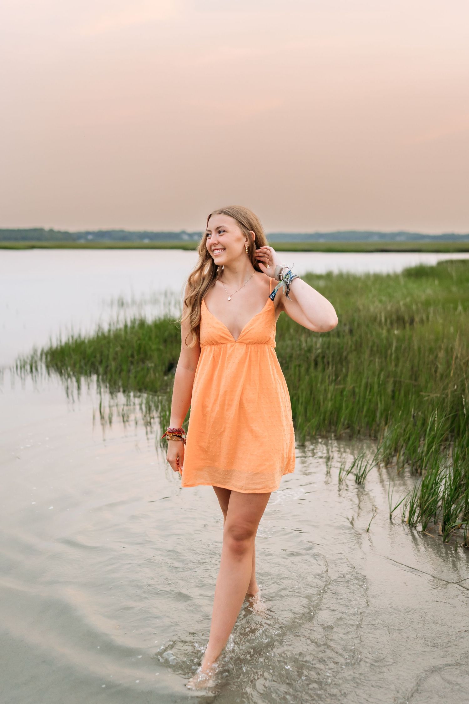 Young woman in orange sundress wading in shallow water near marsh grass at sunset, smiling and touching her hair.