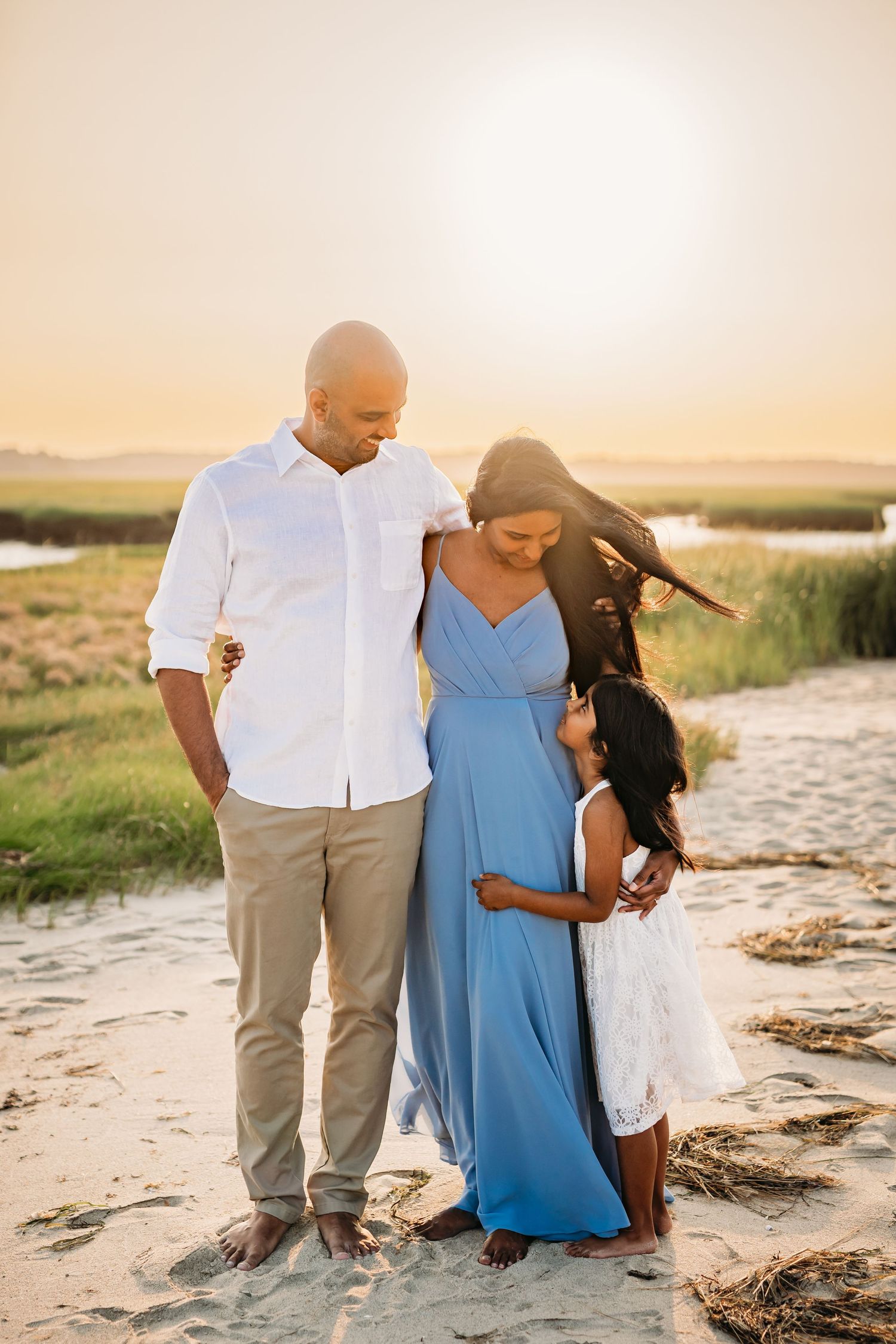 A family in coordinated blue and white outfits walks together along a sandy beach at sunset.