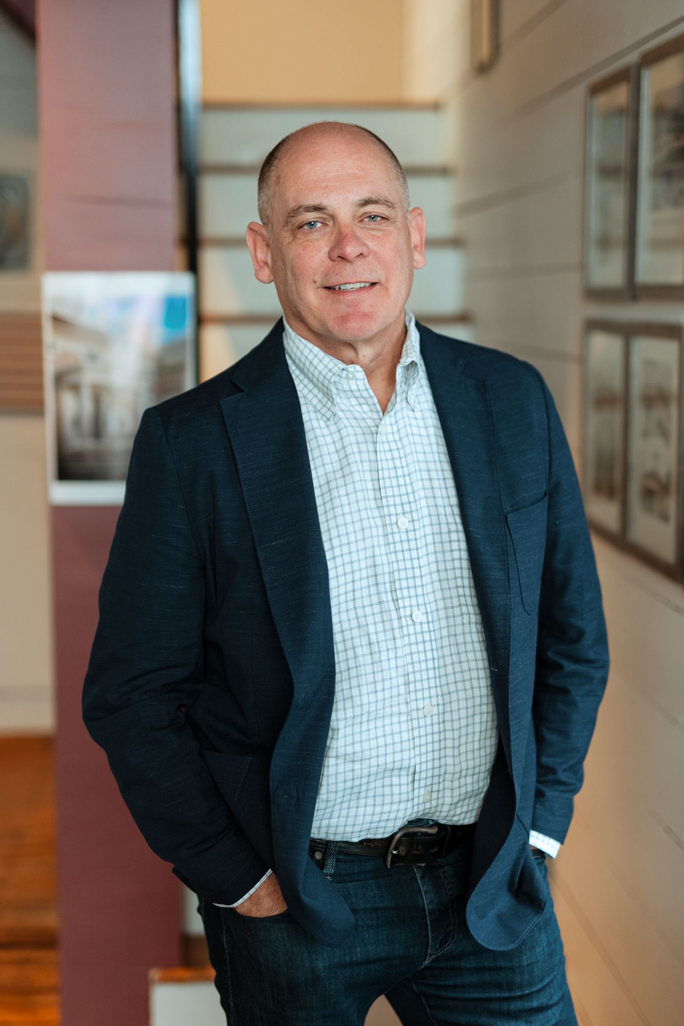Professional man in navy blazer and checkered shirt smiling confidently in a modern office hallway.