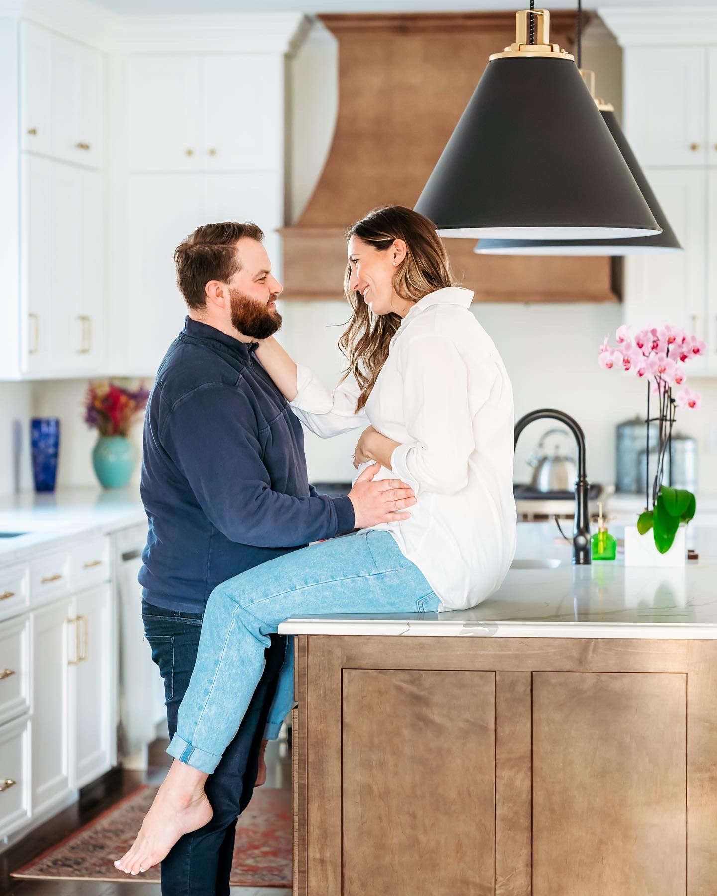 A couple embraces in a modern white kitchen with wooden accents and black pendant lighting.