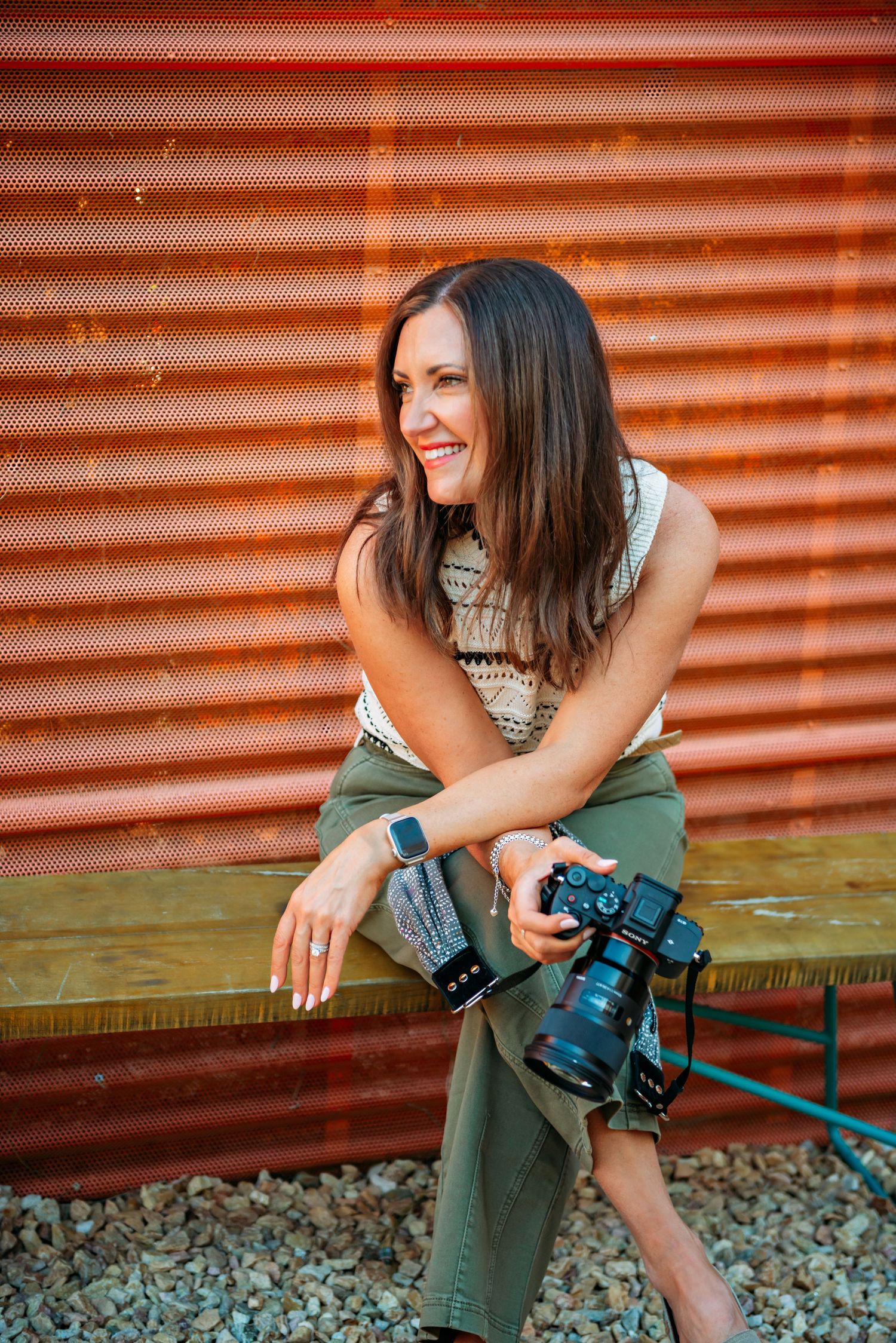 A photographer in green pants sits on a bench with a camera, laughing joyfully against an orange corrugated wall.