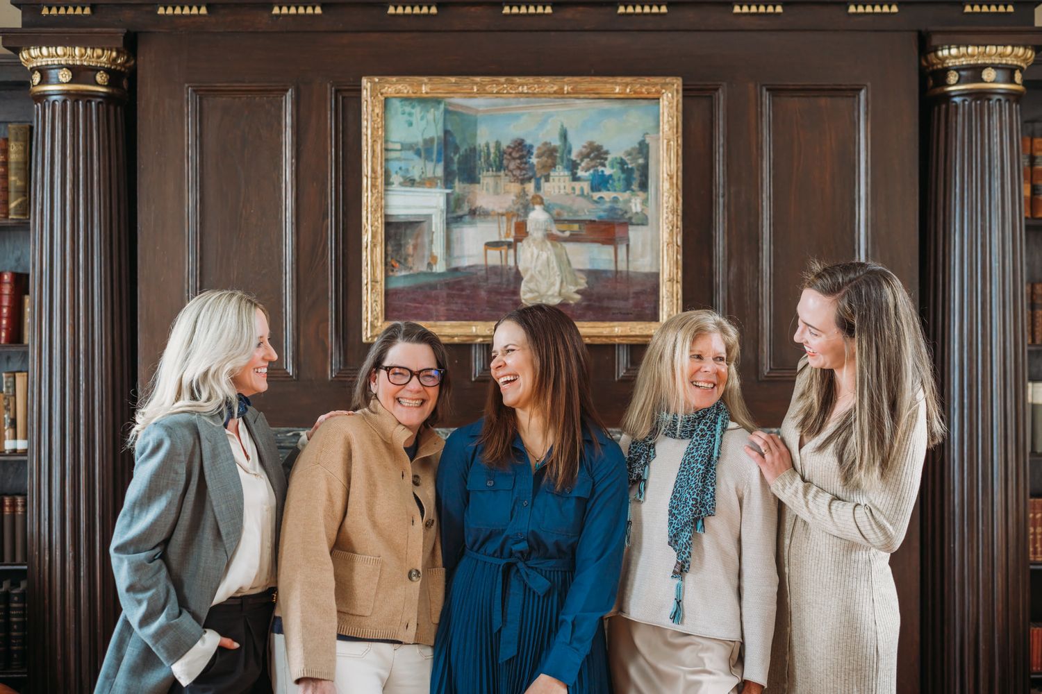Five women smiling and laughing together in an elegant wood-paneled room with a classic painting on the wall.