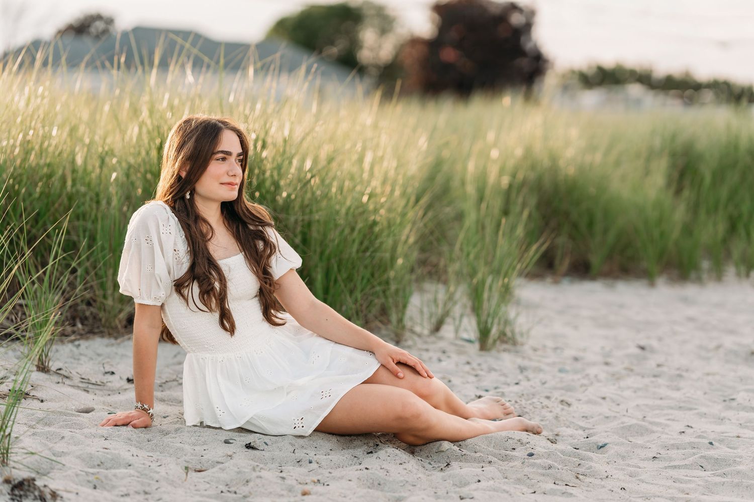 A person in a white dress poses gracefully on a sandy beach with tall grass and water in the background.