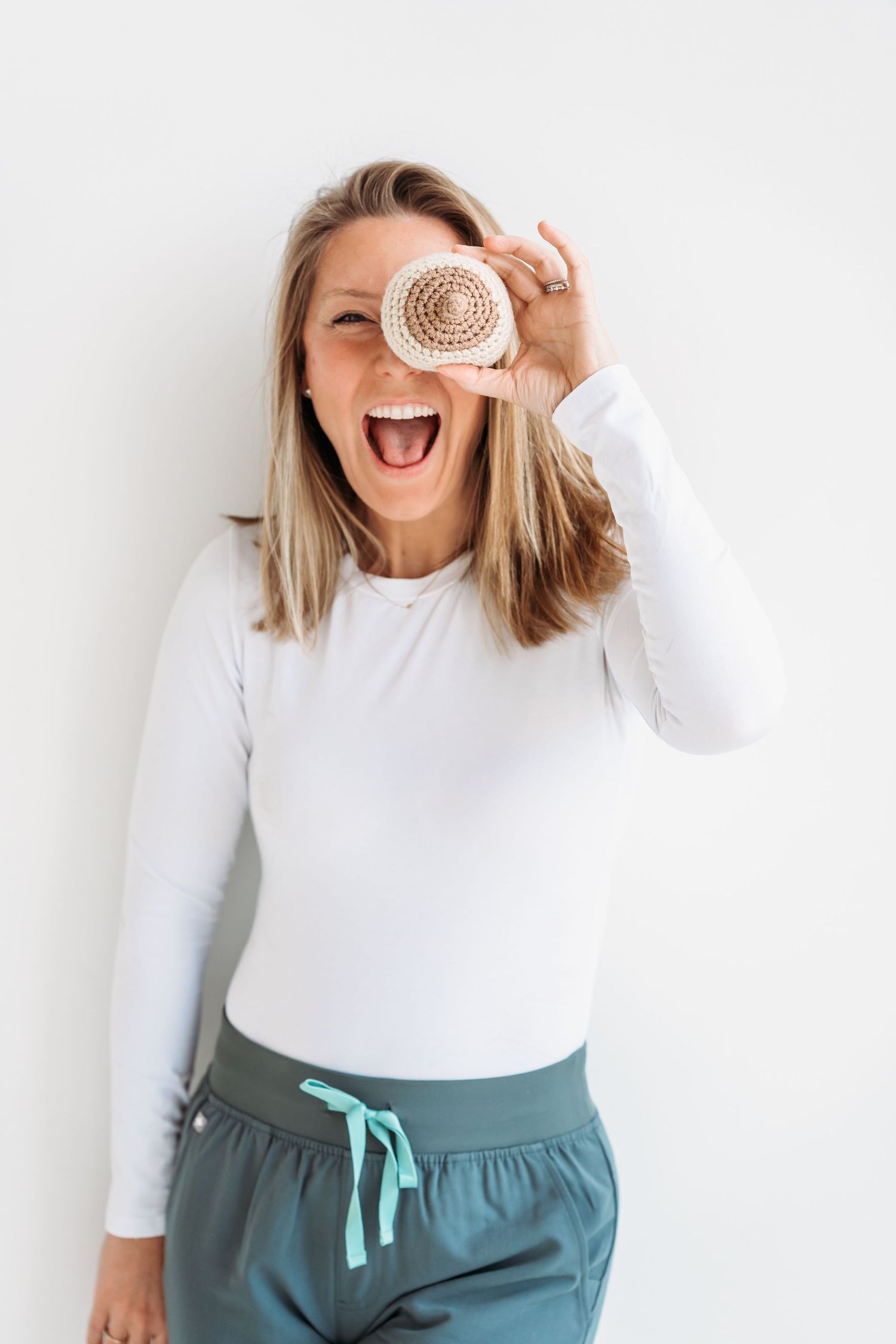 A playful lifestyle photo shows someone in a white top and teal pants holding a donut over their eye while laughing.