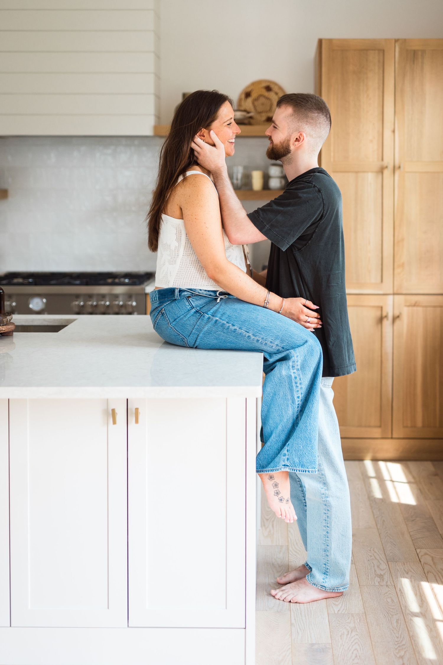 A couple embraces intimately in a bright modern kitchen with white cabinets and wooden floors.