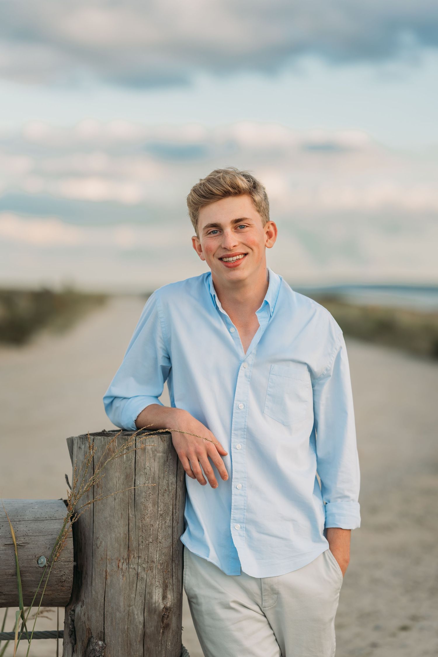 Young man in light blue shirt smiling on sandy beach path, leaning on wooden post with cloudy sky background.