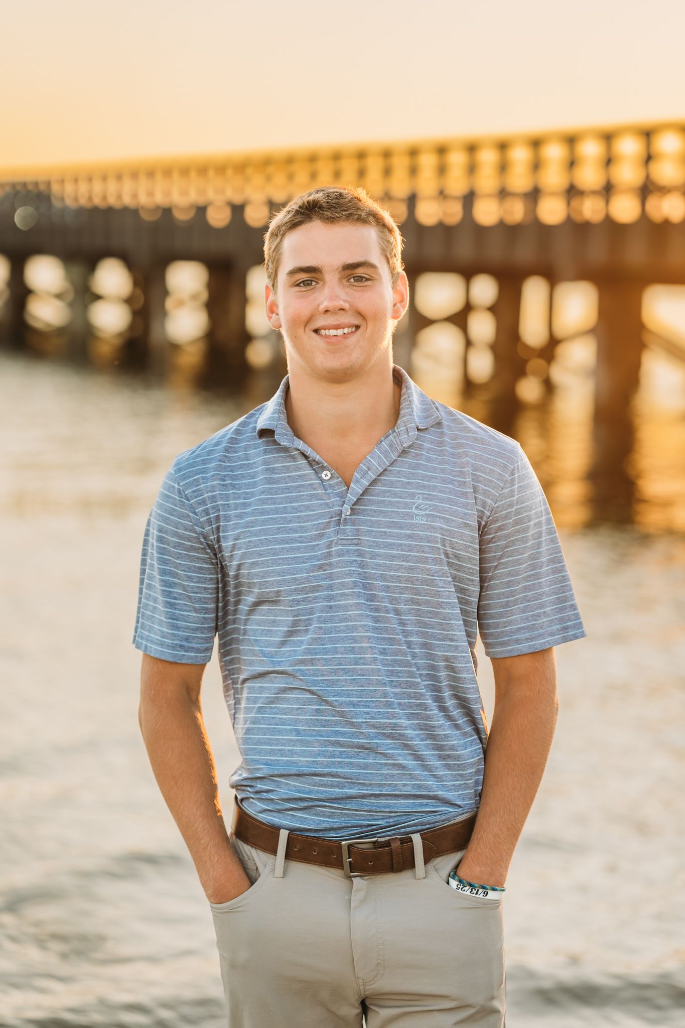 Smiling young man in striped polo shirt standing near a pier at golden hour sunset.