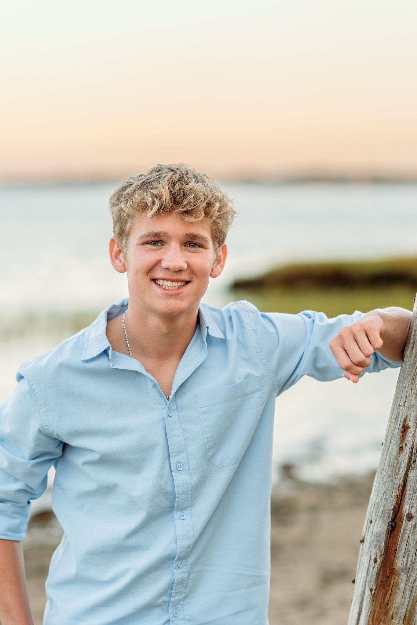 Smiling teenage boy with blond curly hair in light blue shirt leaning on driftwood at beach during sunset.