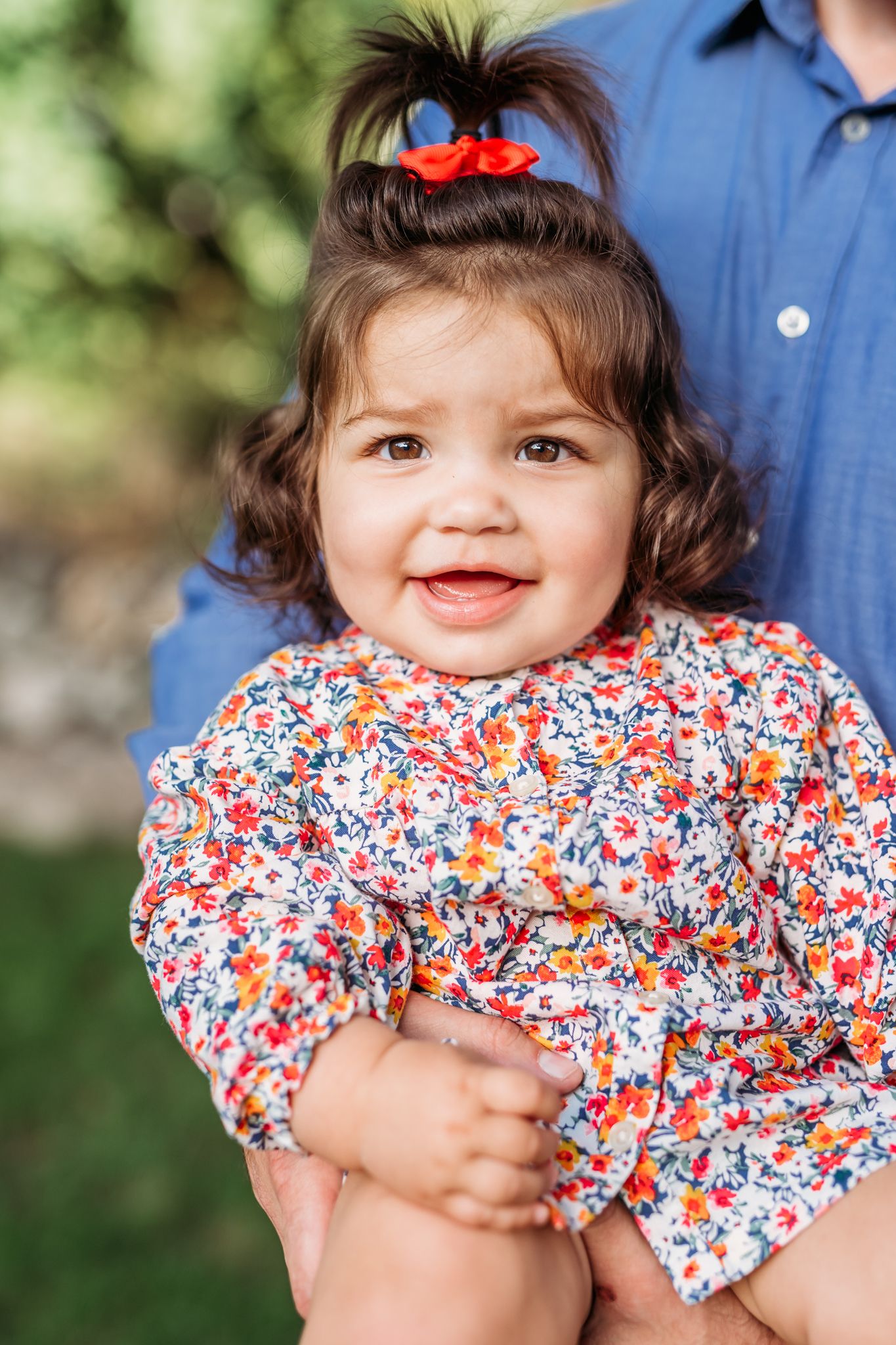 A small child wearing a red, white and blue floral dress smiles while being held outdoors.