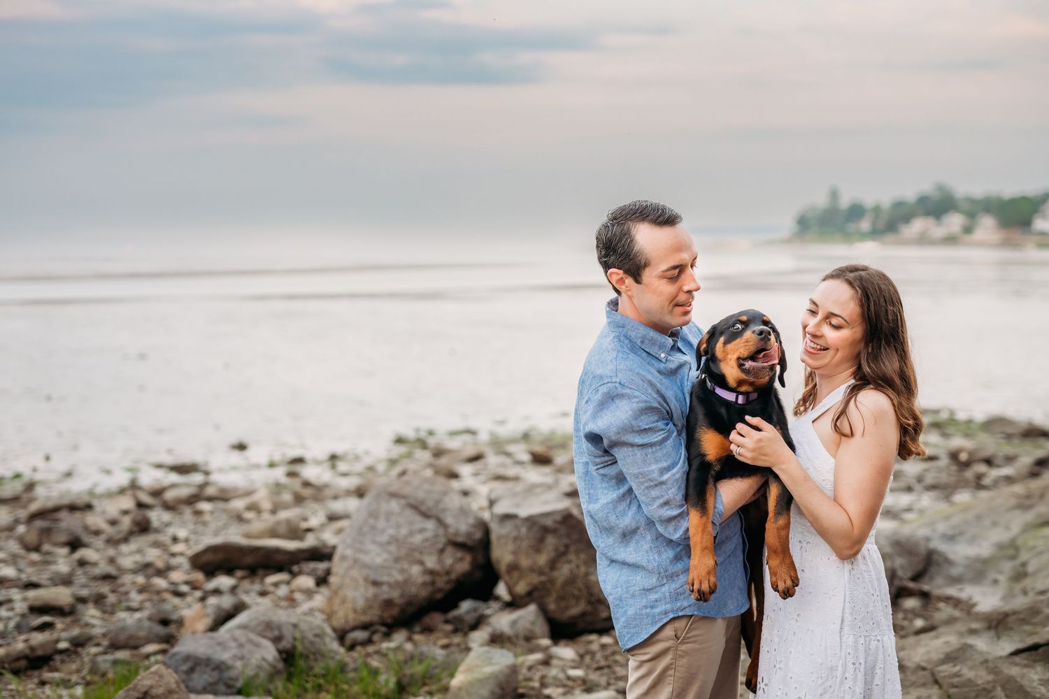 Romantic coastal engagement photo session on a rocky beach during sunset with soft pastel lighting.