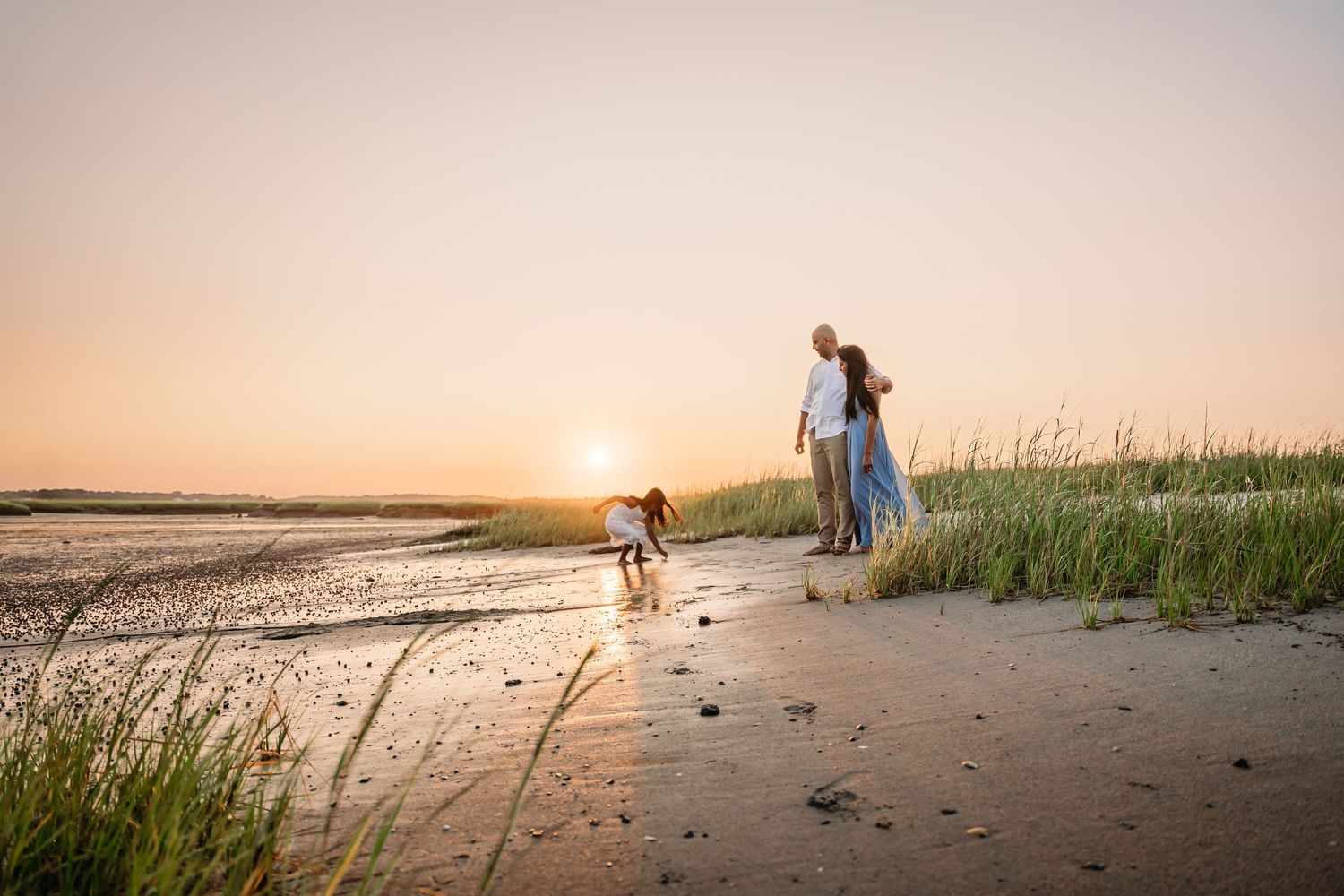 Silhouettes of people walking along a beach at sunset with tall grass and golden sky in the background.
