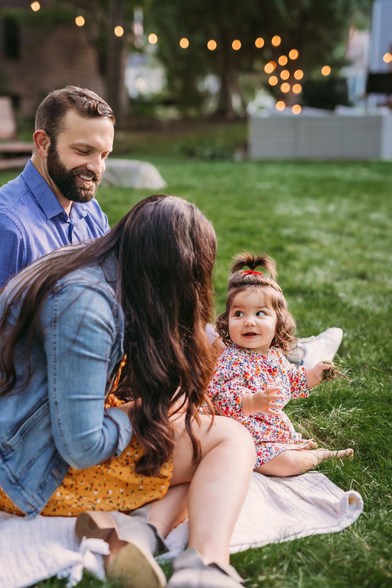 Family enjoying a picnic together on green grass with string lights glowing in the background.