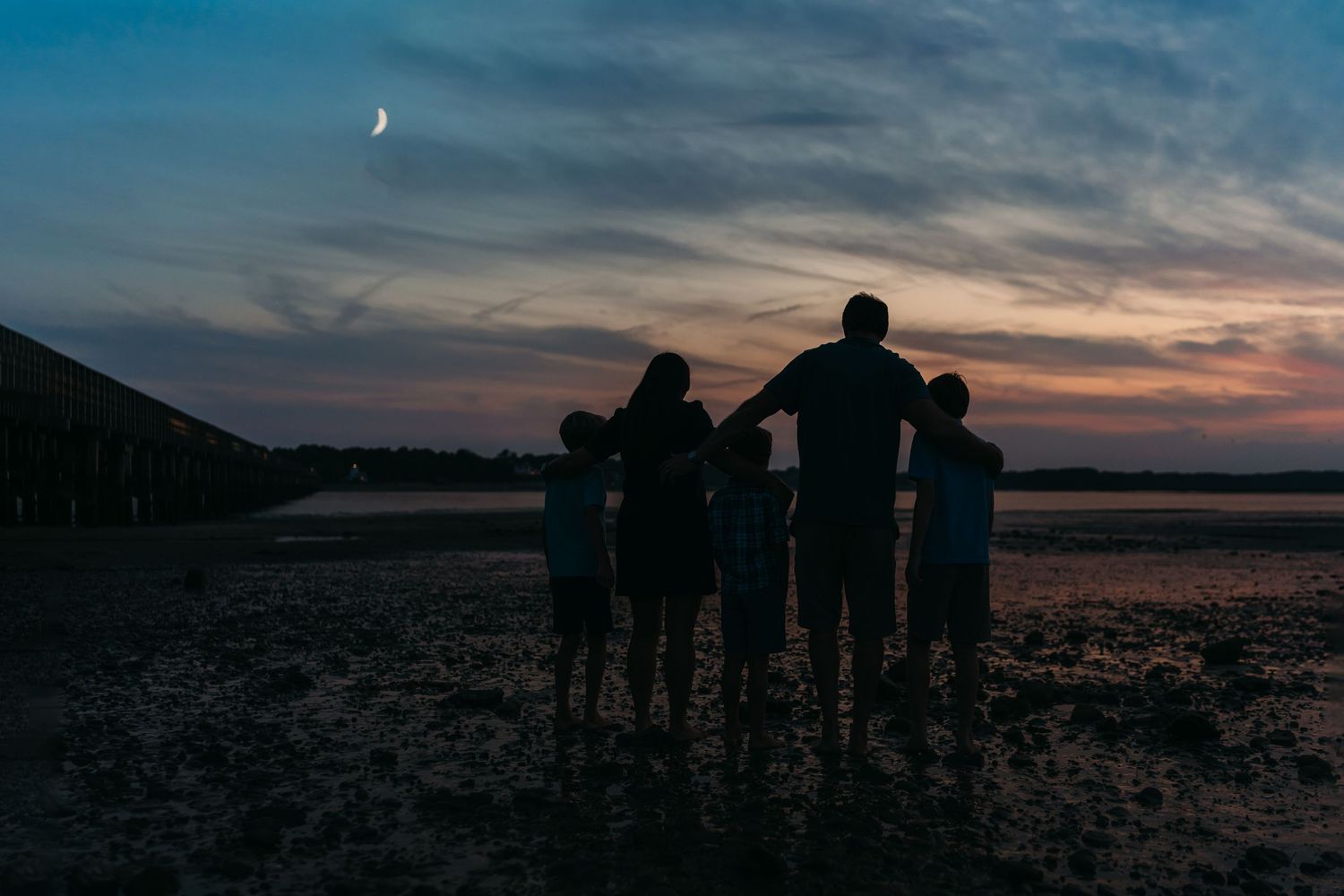A family silhouette stands together watching the moon rise during sunset at a beach landscape.