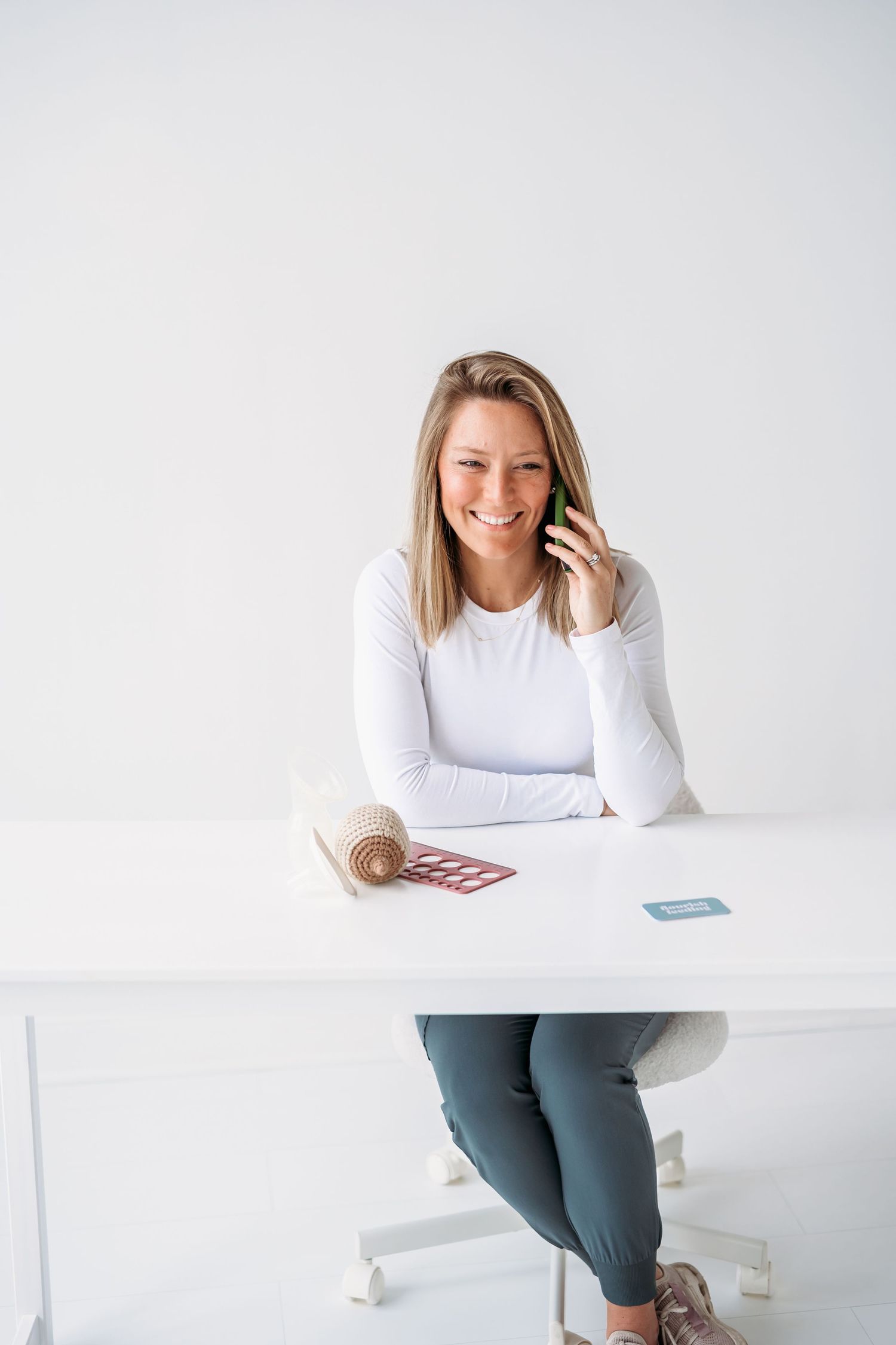 Professional woman in white sweater working at minimalist desk with laptop and phone against white background.