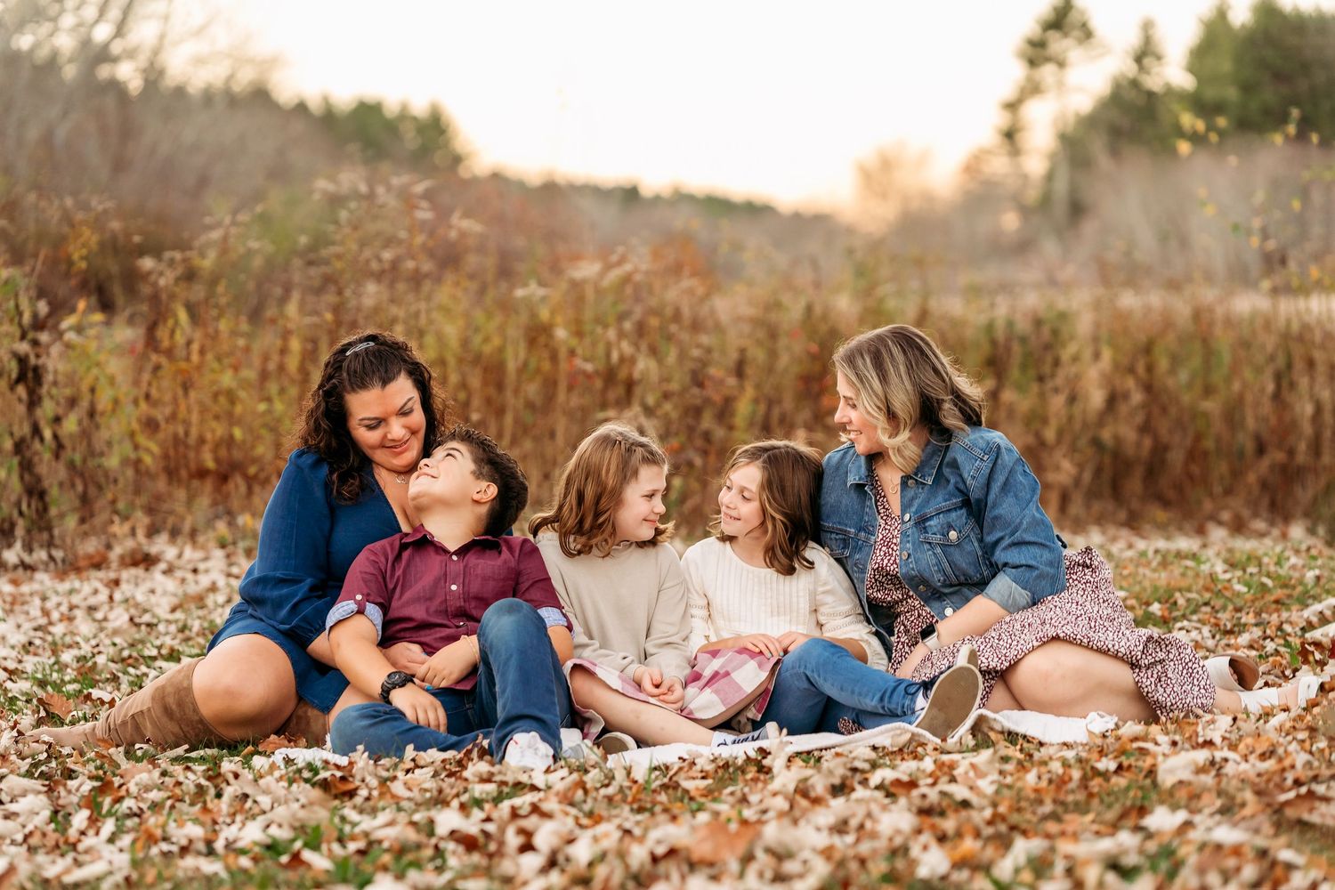 Family members lie together on autumn leaves during an outdoor photo session in a golden field at sunset.