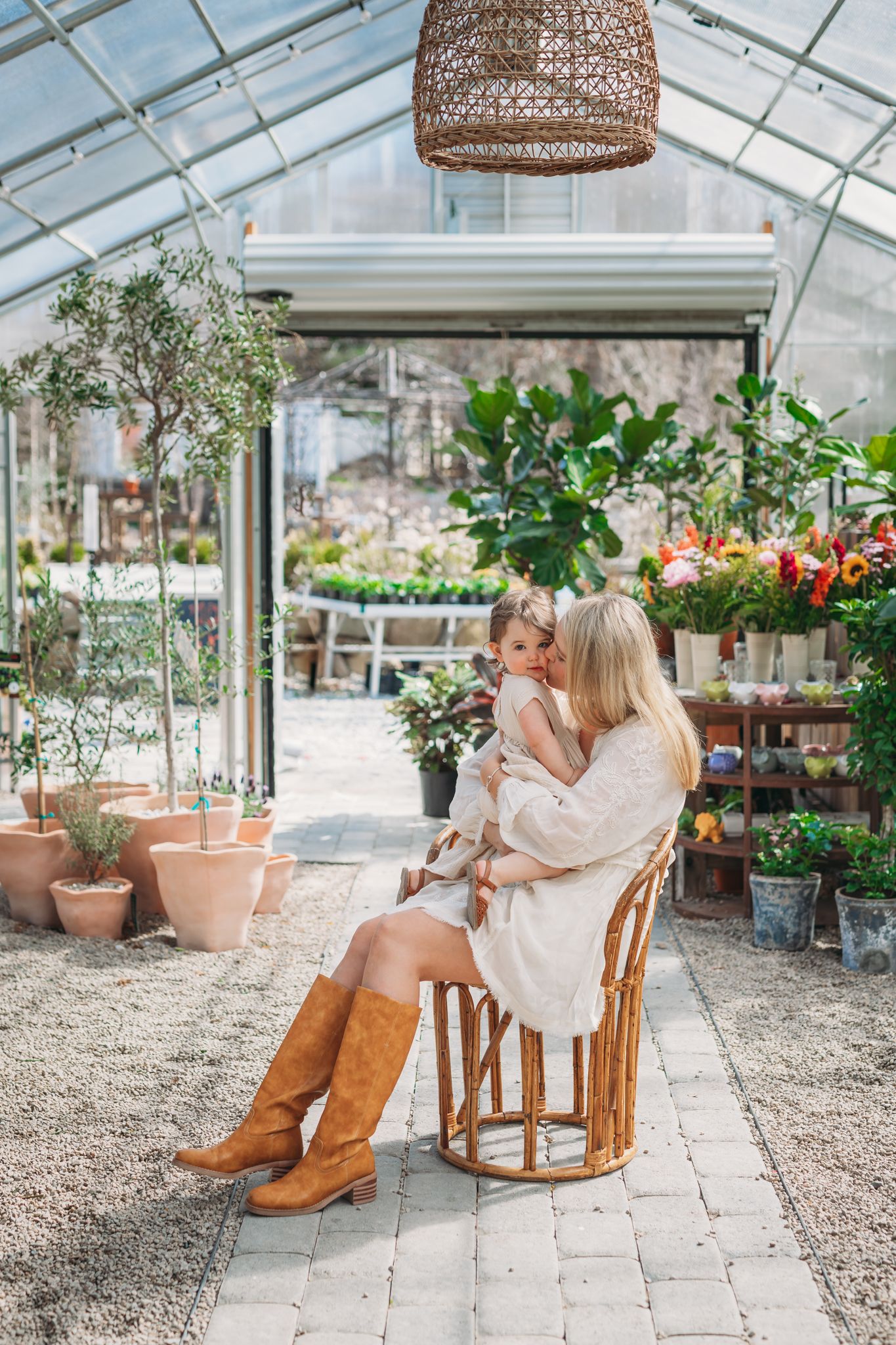 A blonde woman in a cream dress and brown boots sits in a rattan chair holding a toddler in a lush greenhouse.