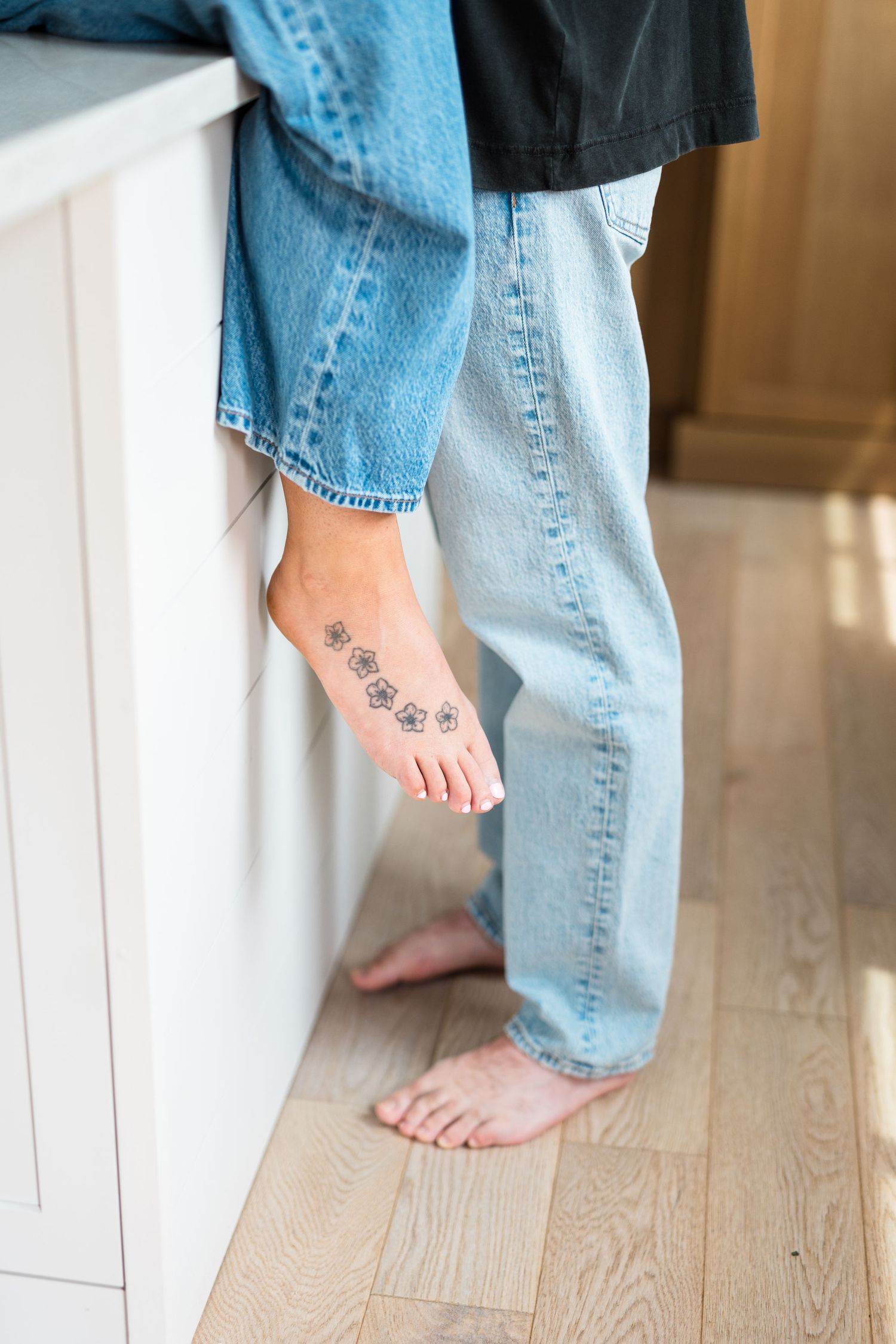Bare feet with small paw print tattoos visible below light blue denim jeans on a hardwood floor.
