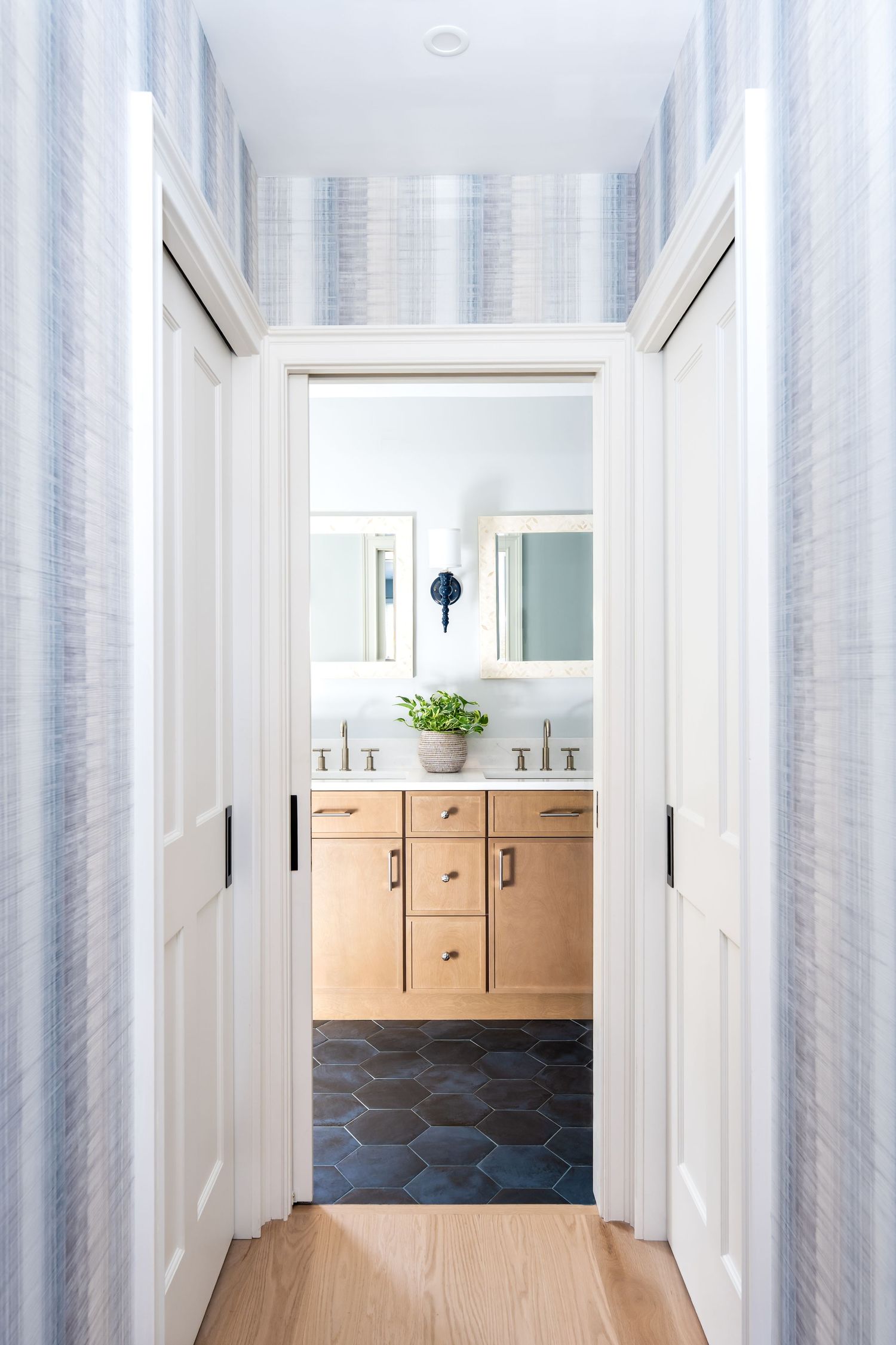 Modern bathroom entrance with light wood vanity, hexagonal black floor tiles, and white walls featuring blue wallpaper.