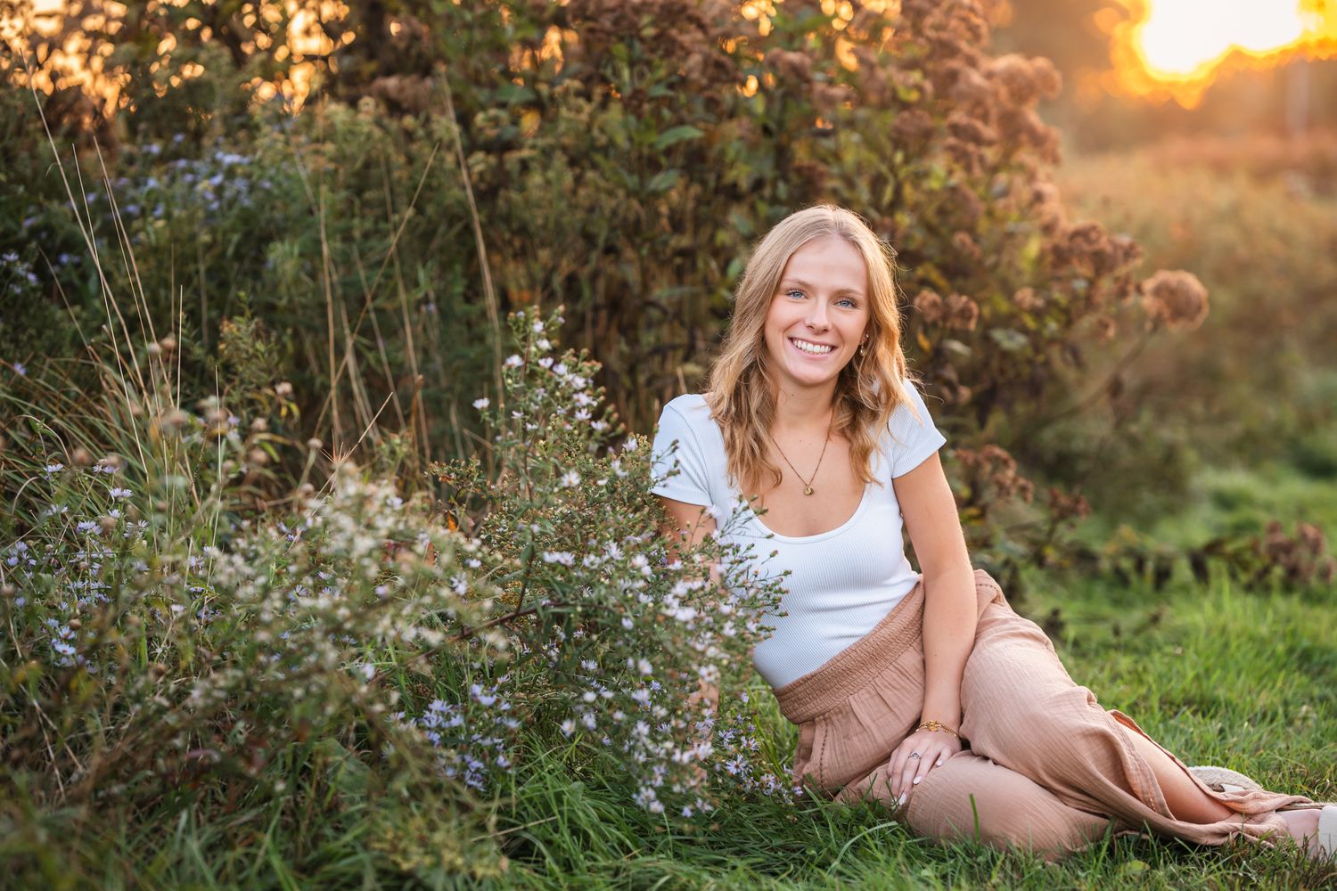 Smiling blonde woman sitting on grass near wildflowers in golden hour sunlight outdoors.