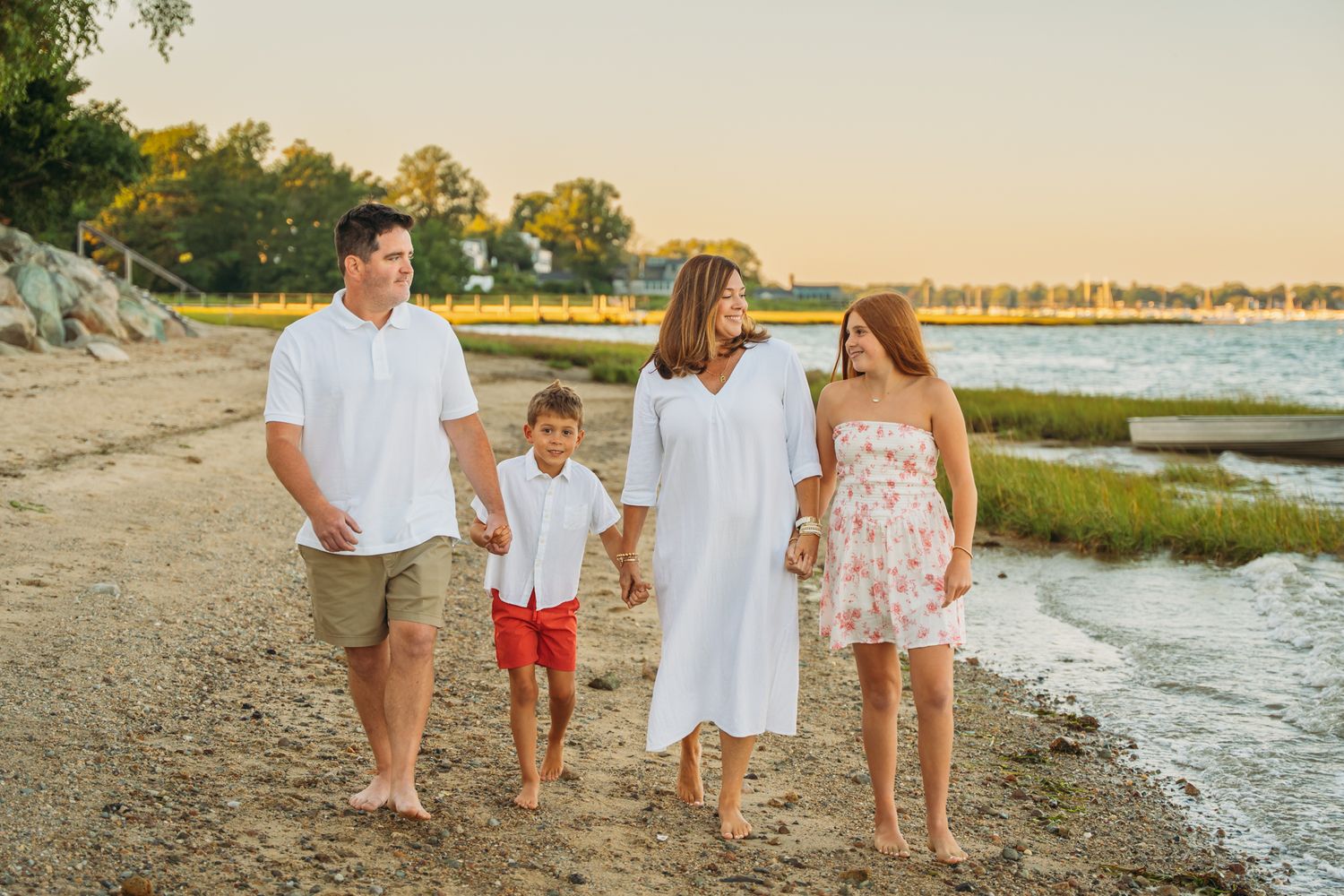 Family of four walking barefoot on a sandy beach at golden hour, wearing white and red outfits near calm water.