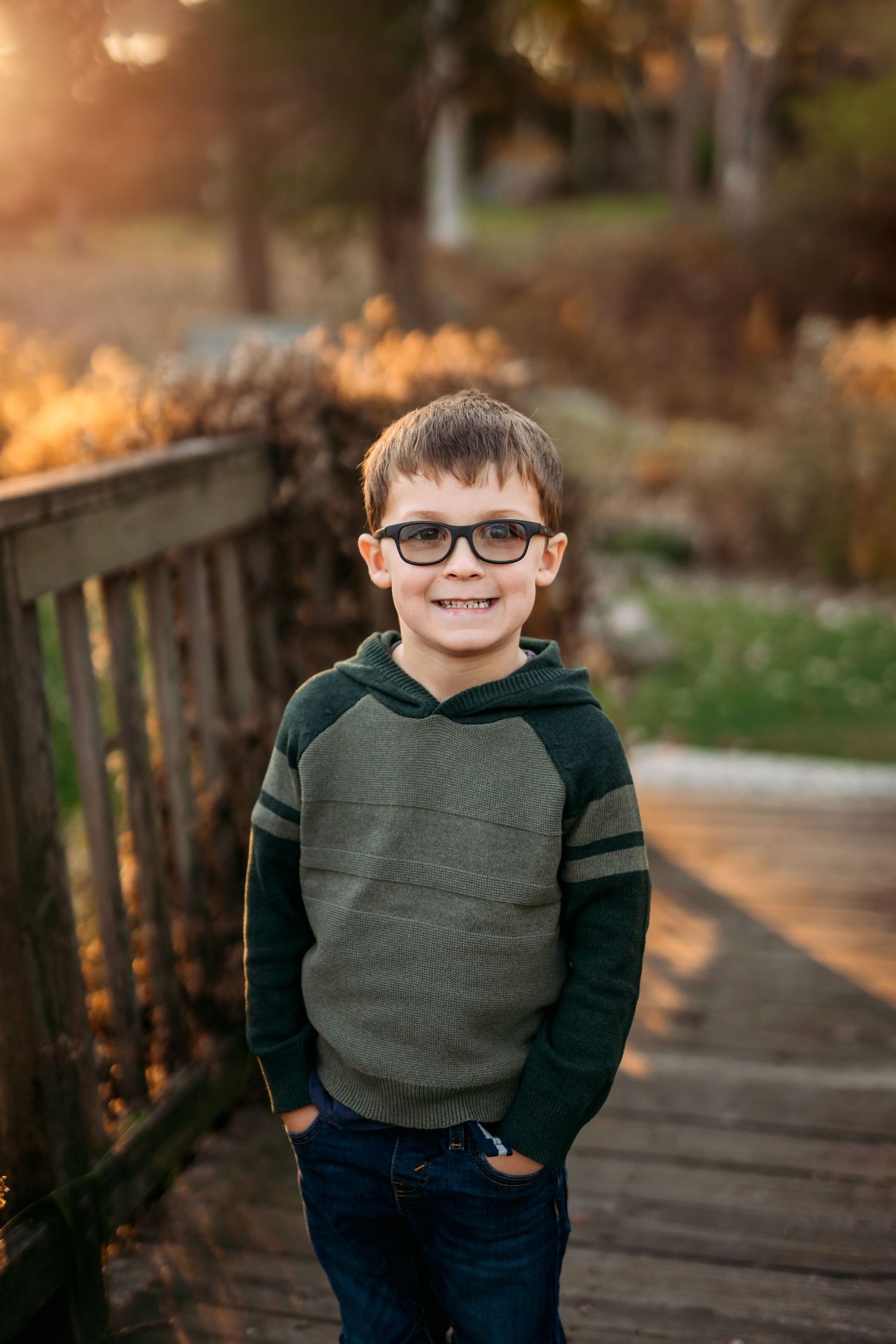 Young child in glasses and green sweater poses on wooden bridge at sunset.