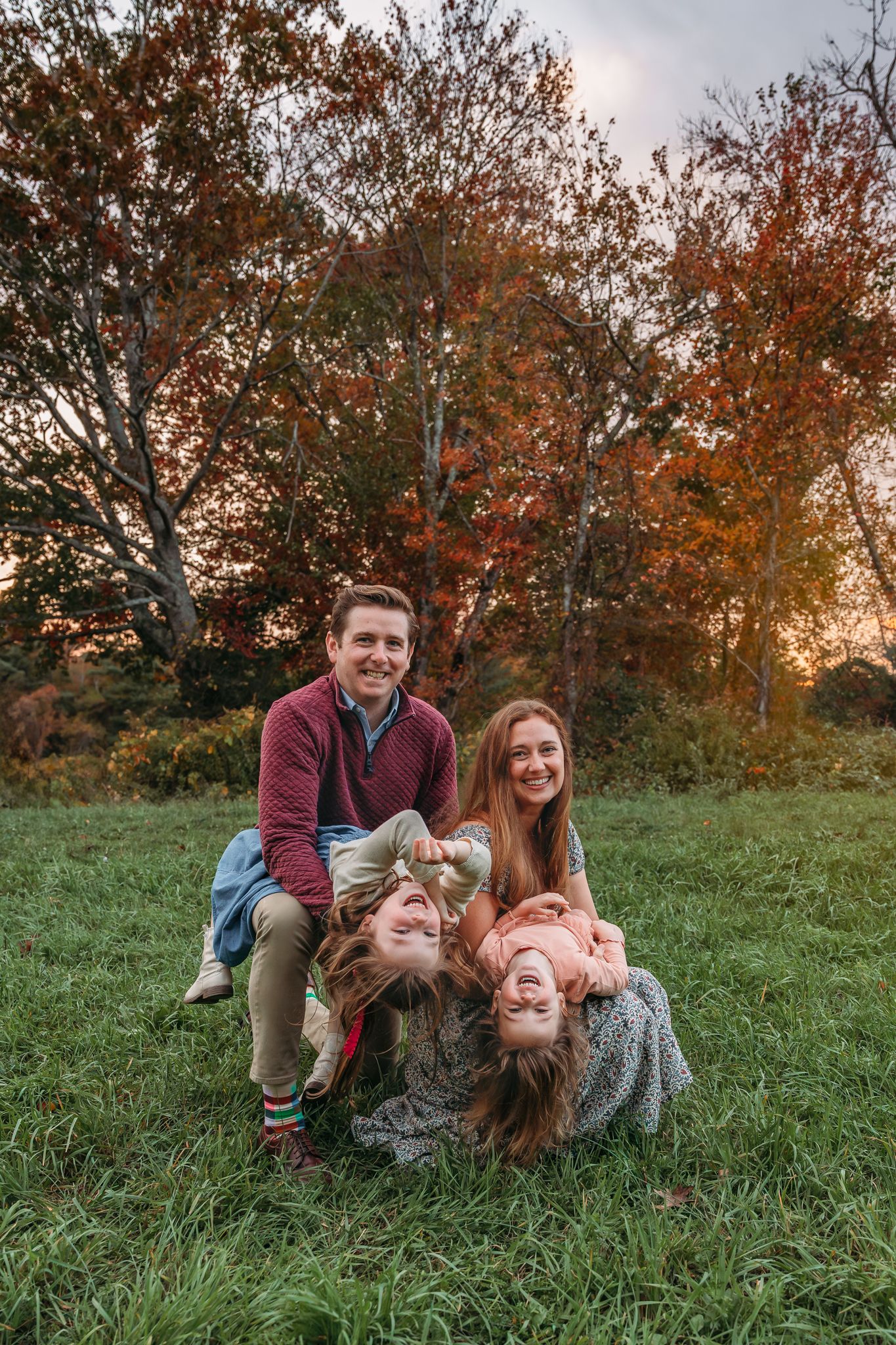 Family of four posing playfully on grass with autumn trees at sunset, parents holding children upside down.