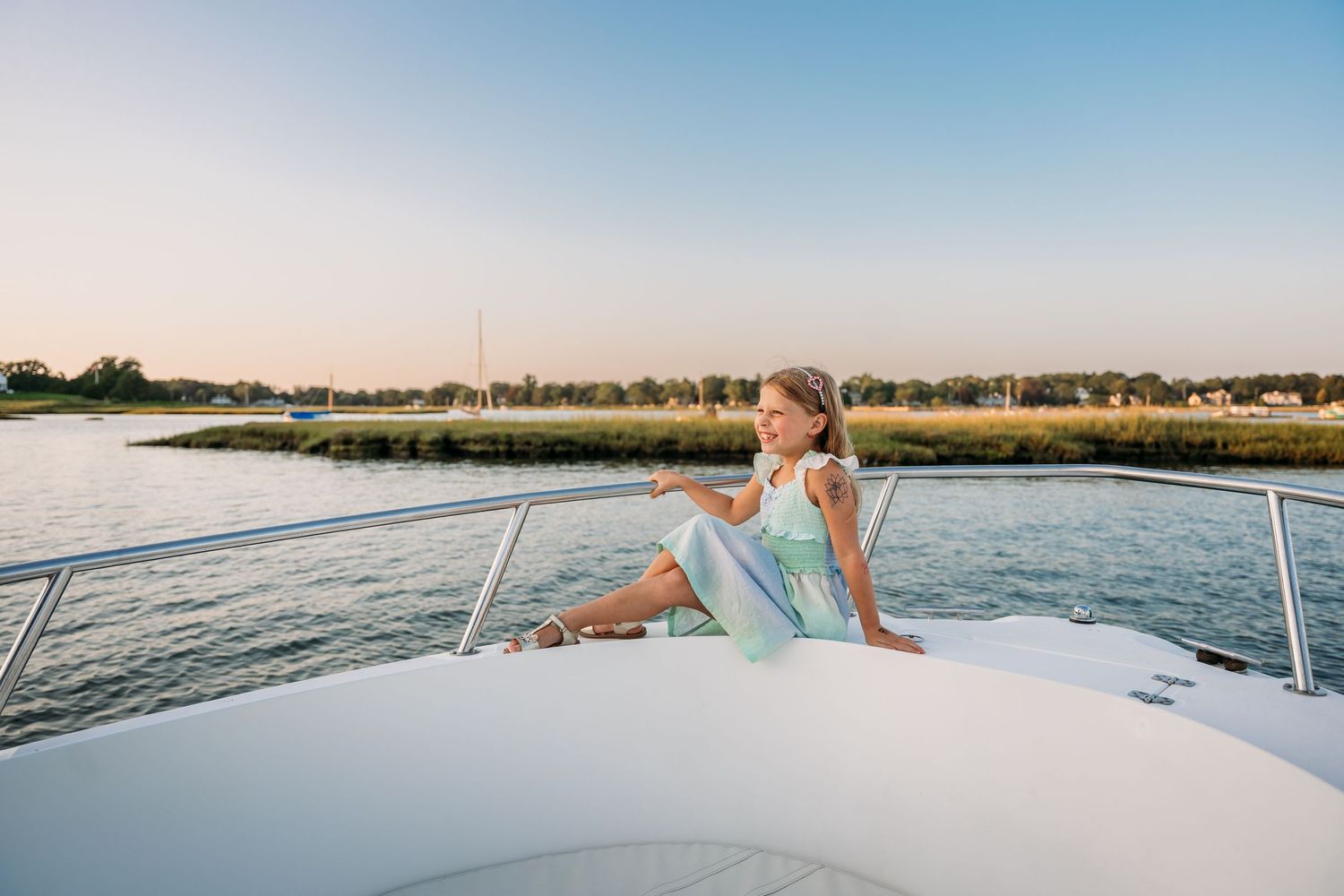 Someone wearing a blue sundress relaxes on the bow of a yacht while cruising along a scenic waterfront at sunset.