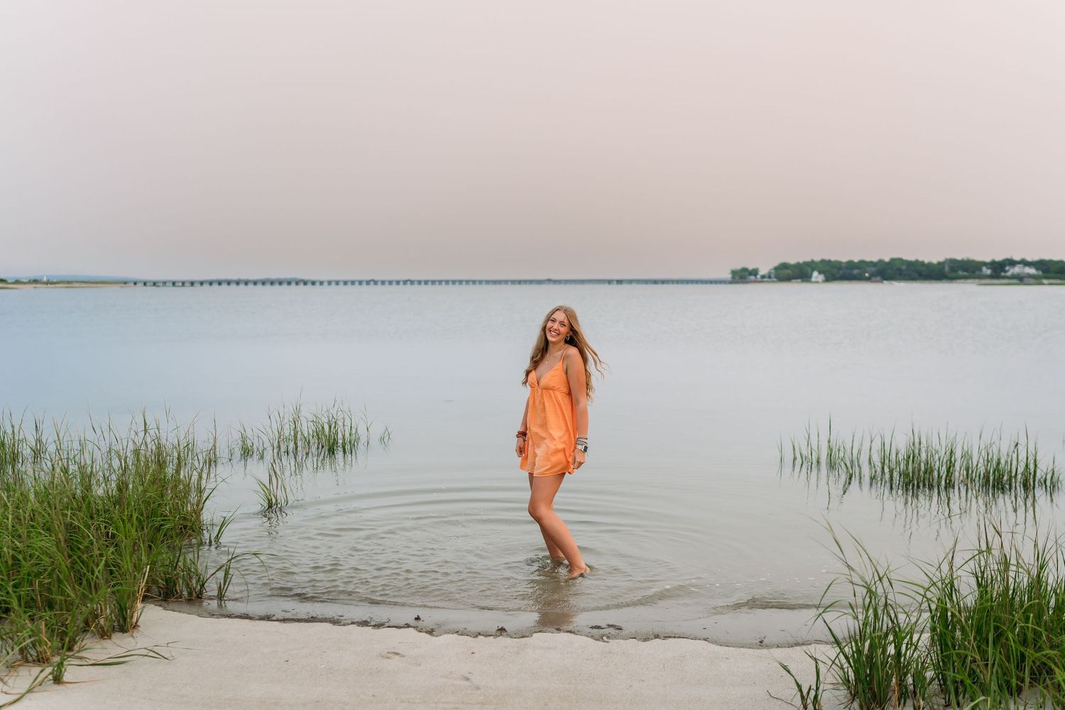 Young woman in orange dress wading in calm coastal waters at dusk, with marsh grass and pier in background.