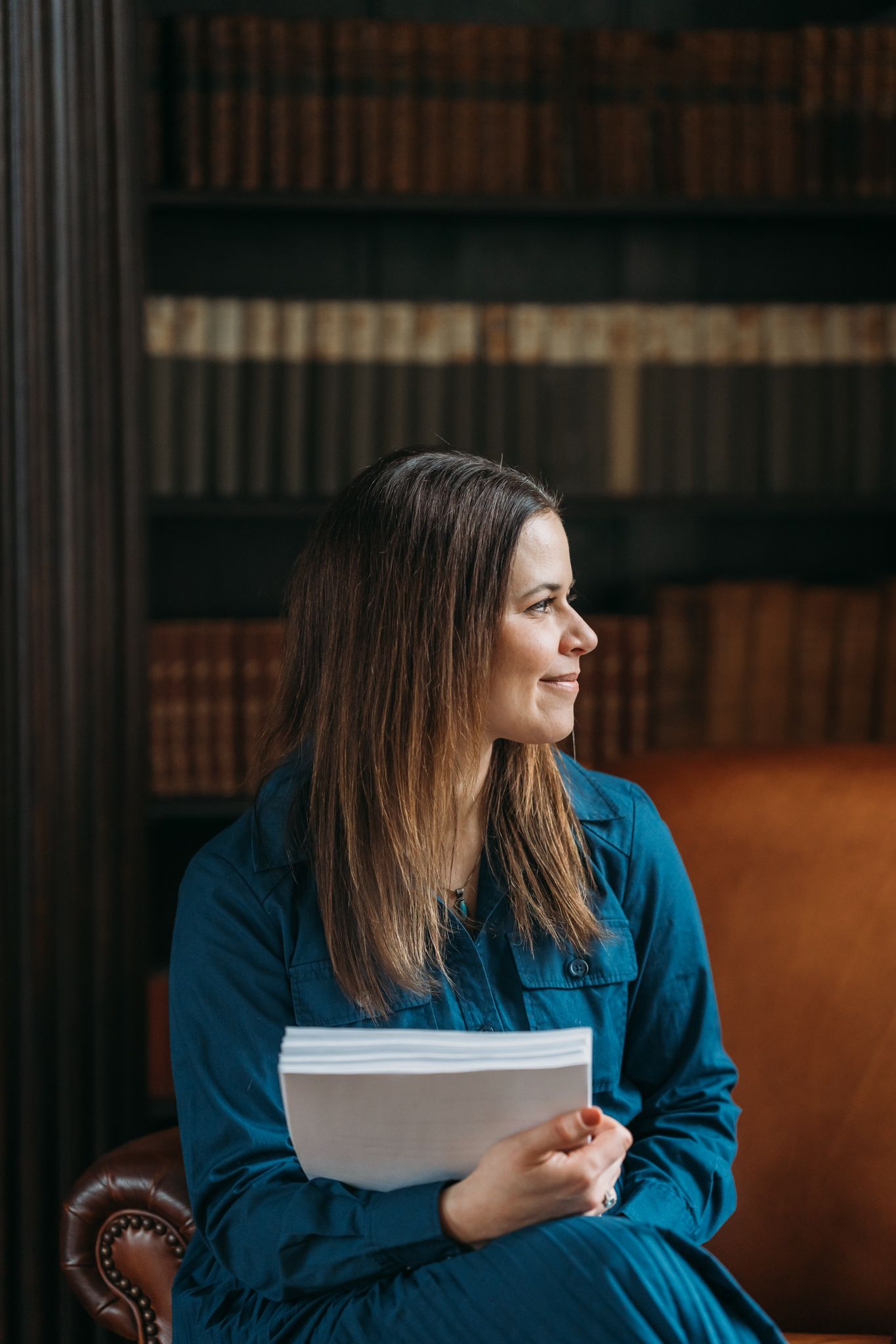 Woman in teal jacket sits in leather chair holding papers, smiling, with vintage bookshelf library background.