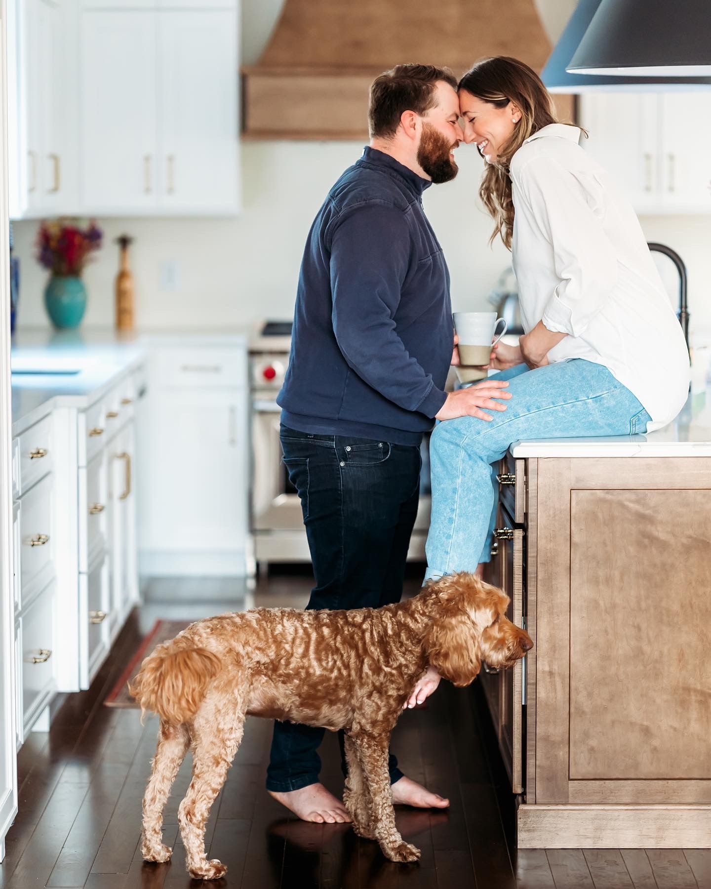A couple shares an intimate moment in their modern kitchen while their golden retriever stands nearby watching them.