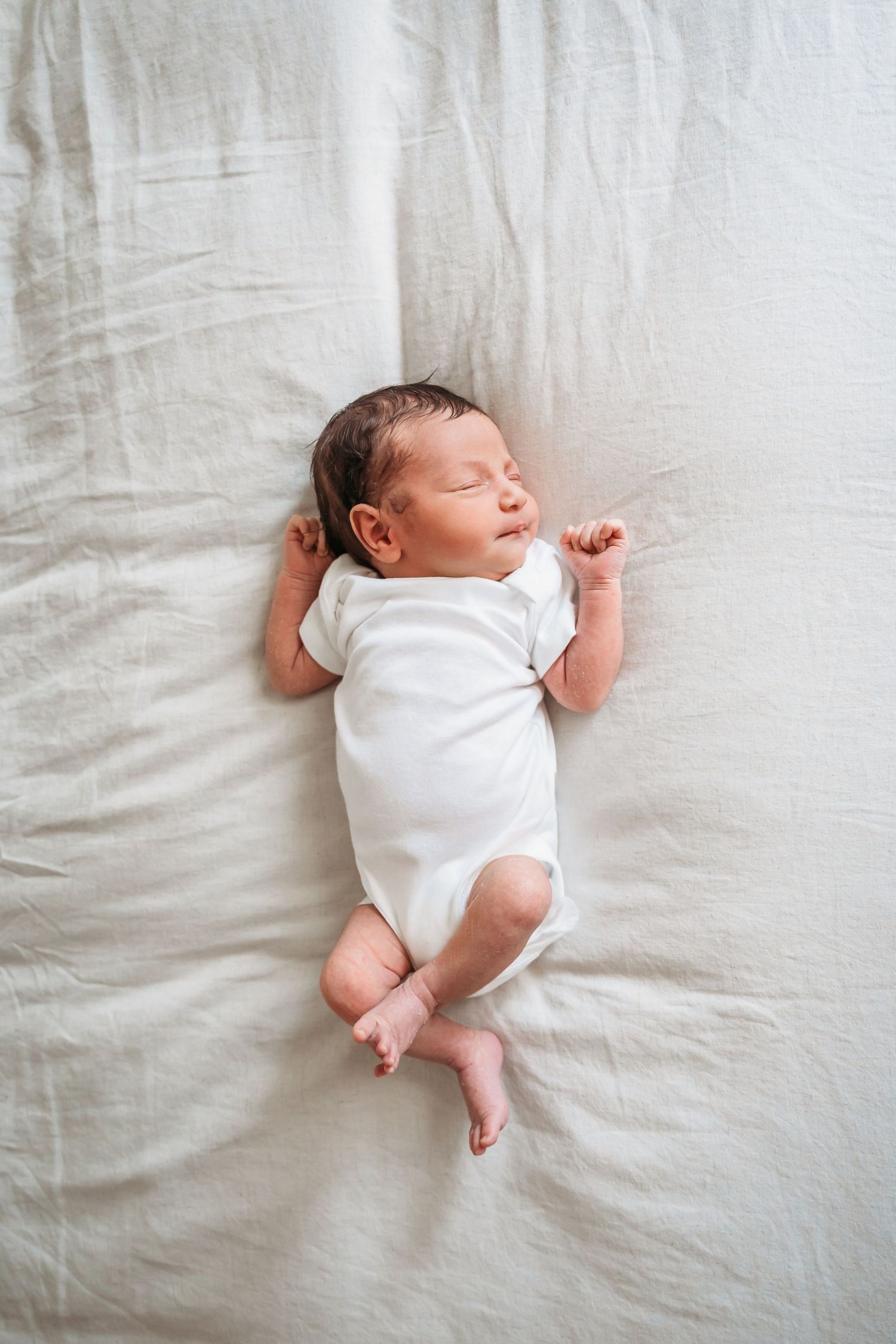 Sleeping newborn baby wearing white onesie lies on neutral linen background.