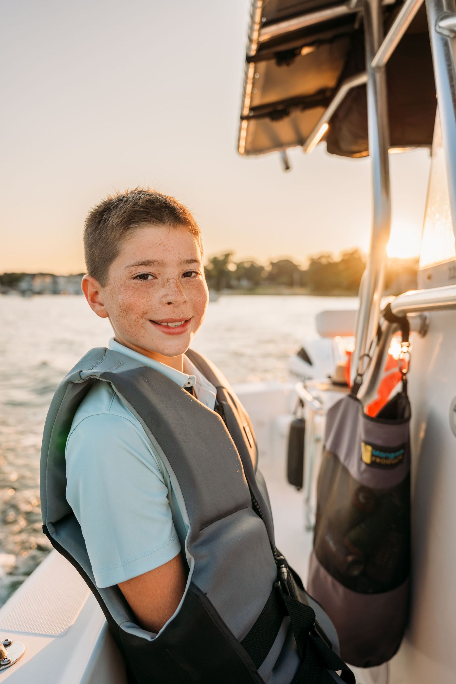 Young person smiling while wearing a gray life vest on a boat during sunset.