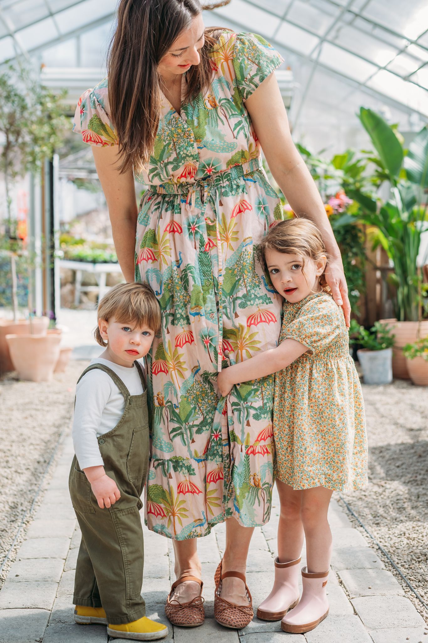 A mother in a floral dress stands with two young children in a bright greenhouse setting.